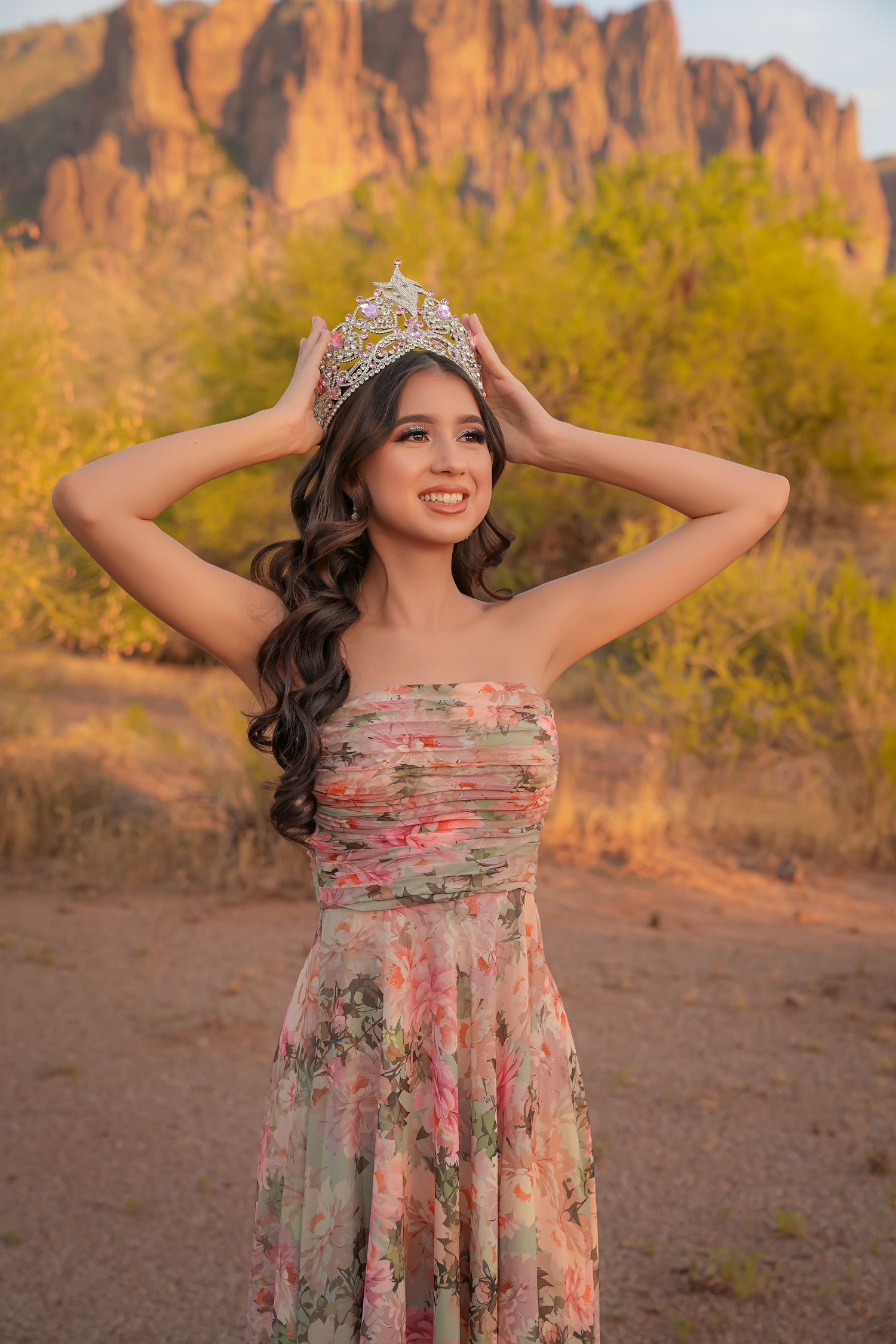 Young woman in floral dress and tiara crown poses outdoors with desert mountains at golden hour.