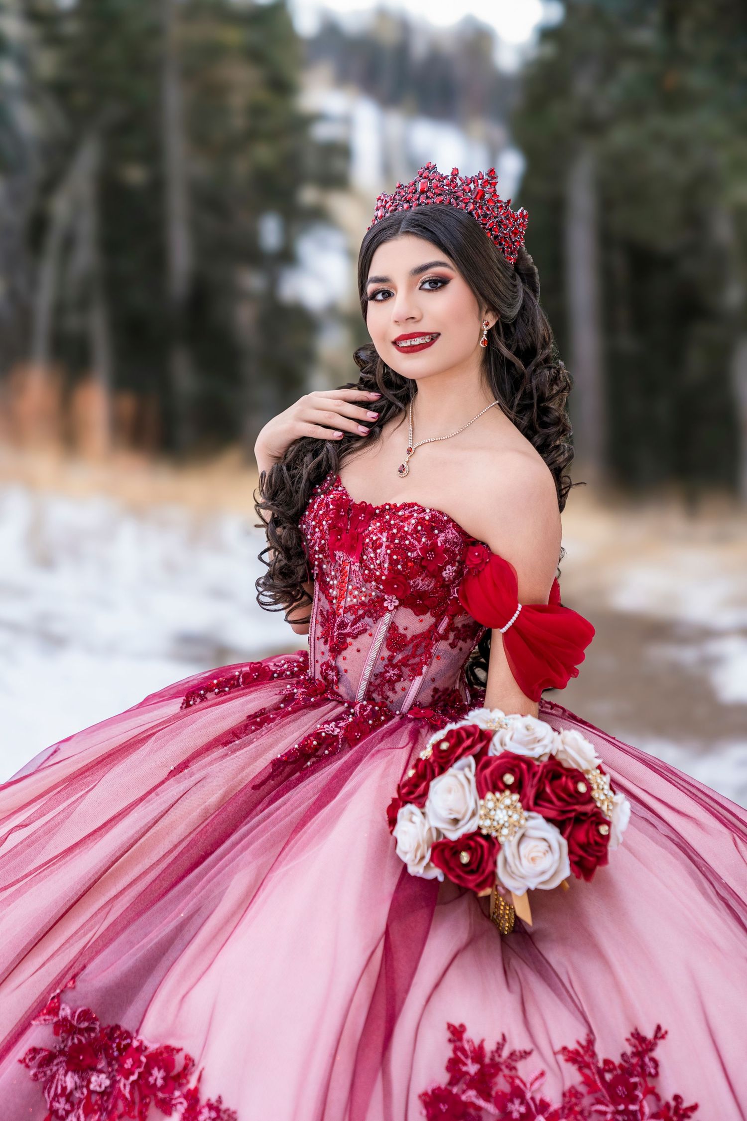 Young woman in red and pink quinceañera ball gown with crown holds bouquet in a snowy winter setting.