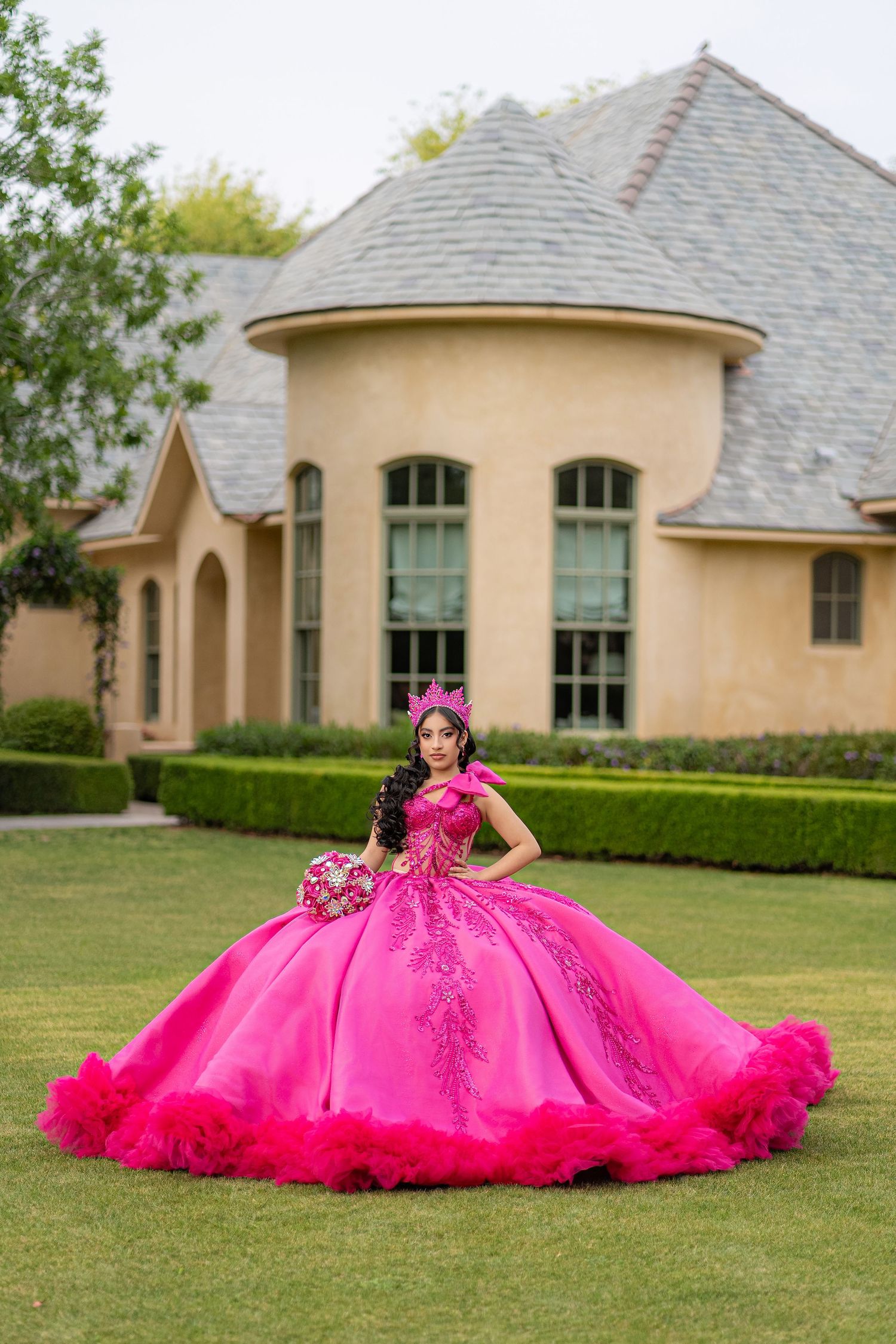 A young woman poses in a vibrant hot pink quinceañera ball gown with floral details and crown on a manicured lawn.