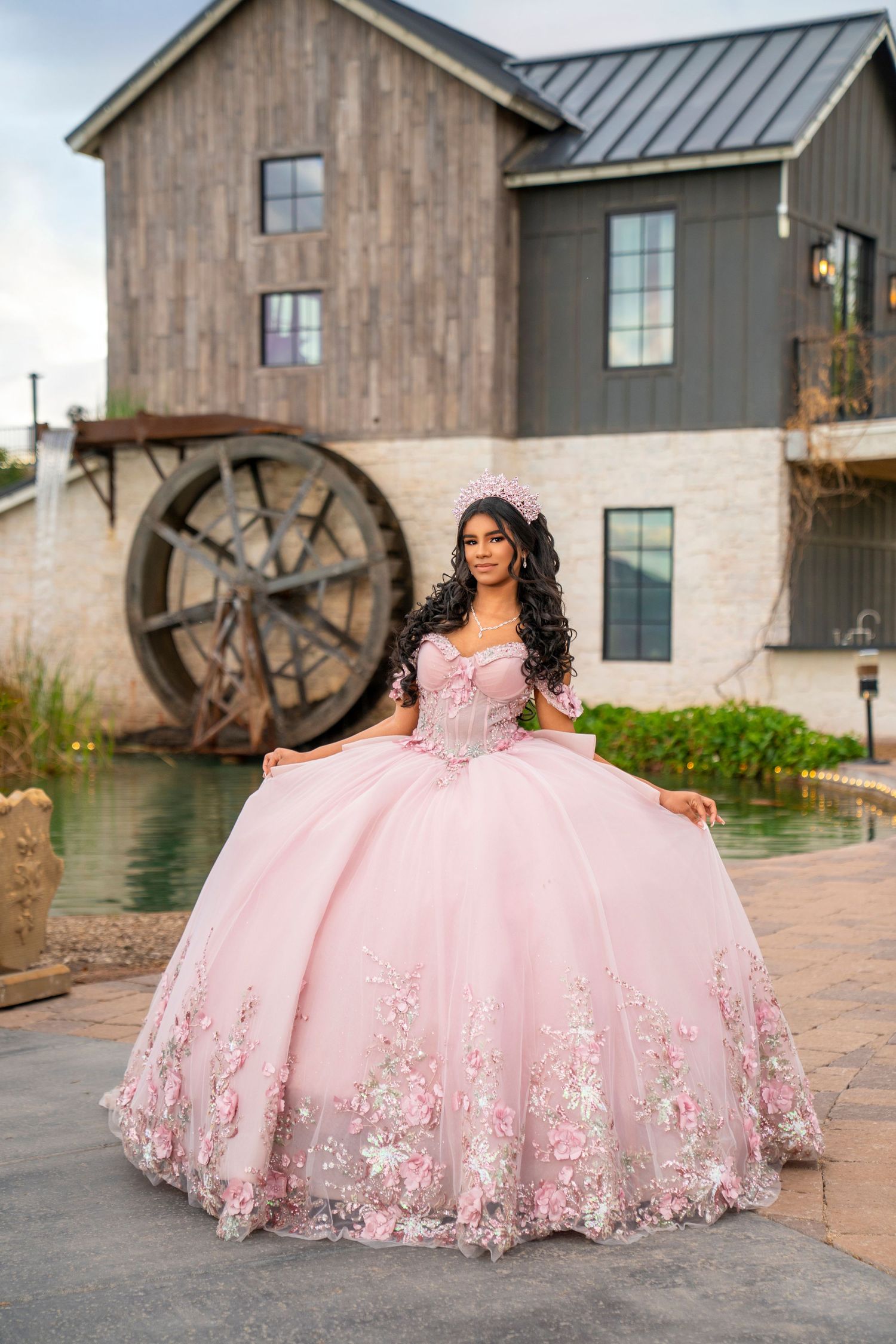 Young woman in pink quinceañera ball gown with floral details poses near rustic mill building.