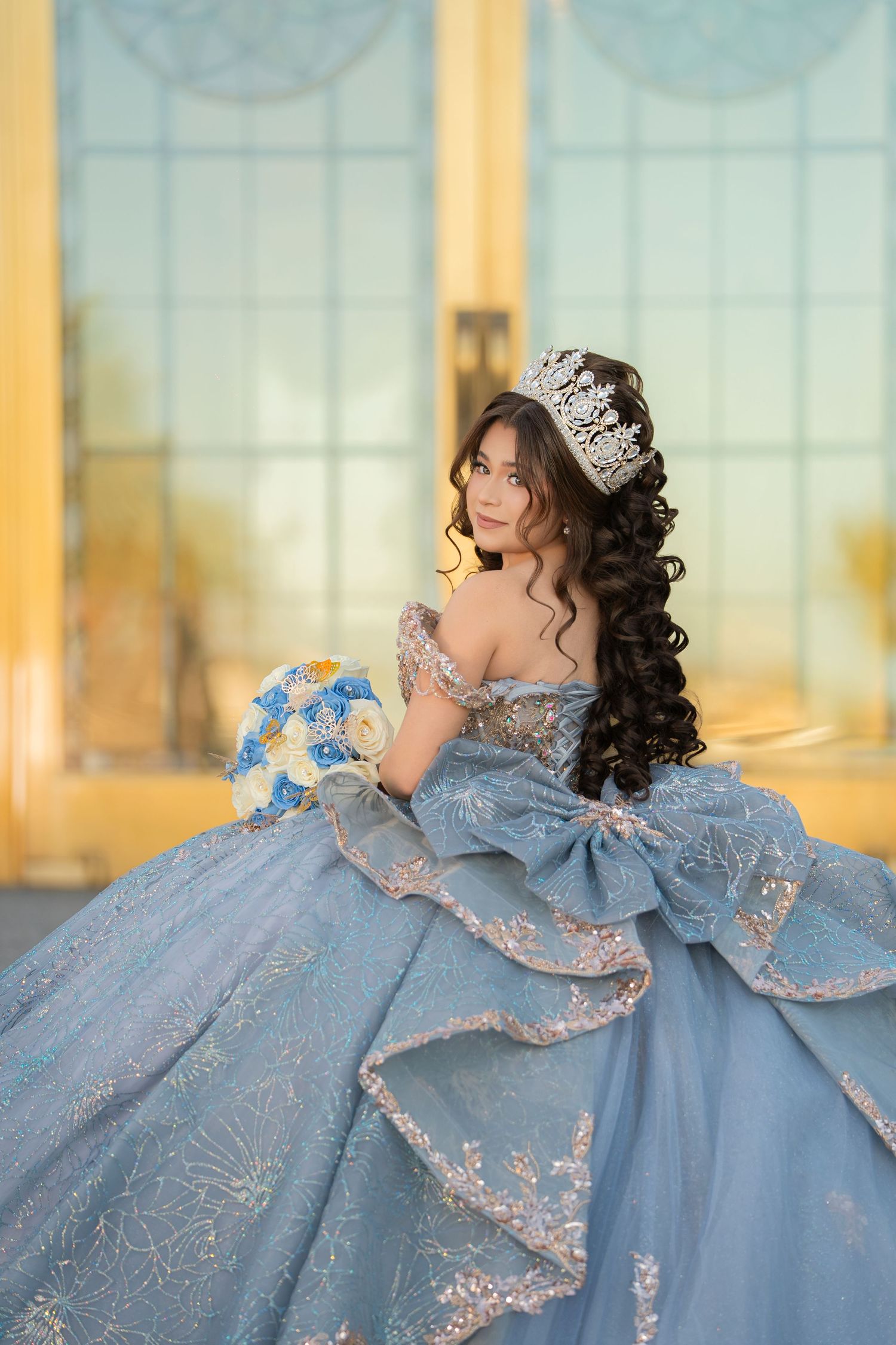 Young woman in a blue ball gown with crown and floral bouquet poses elegantly for a quinceañera celebration.