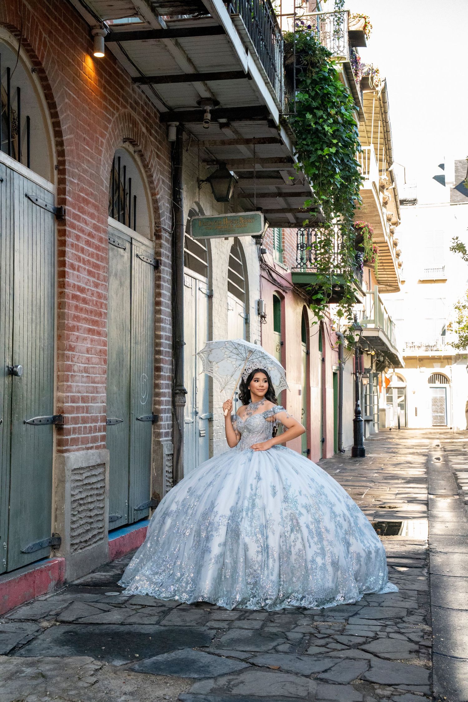 Young woman in a white ball gown quinceañera dress holding a lace umbrella on a historic New Orleans French Quarter street.