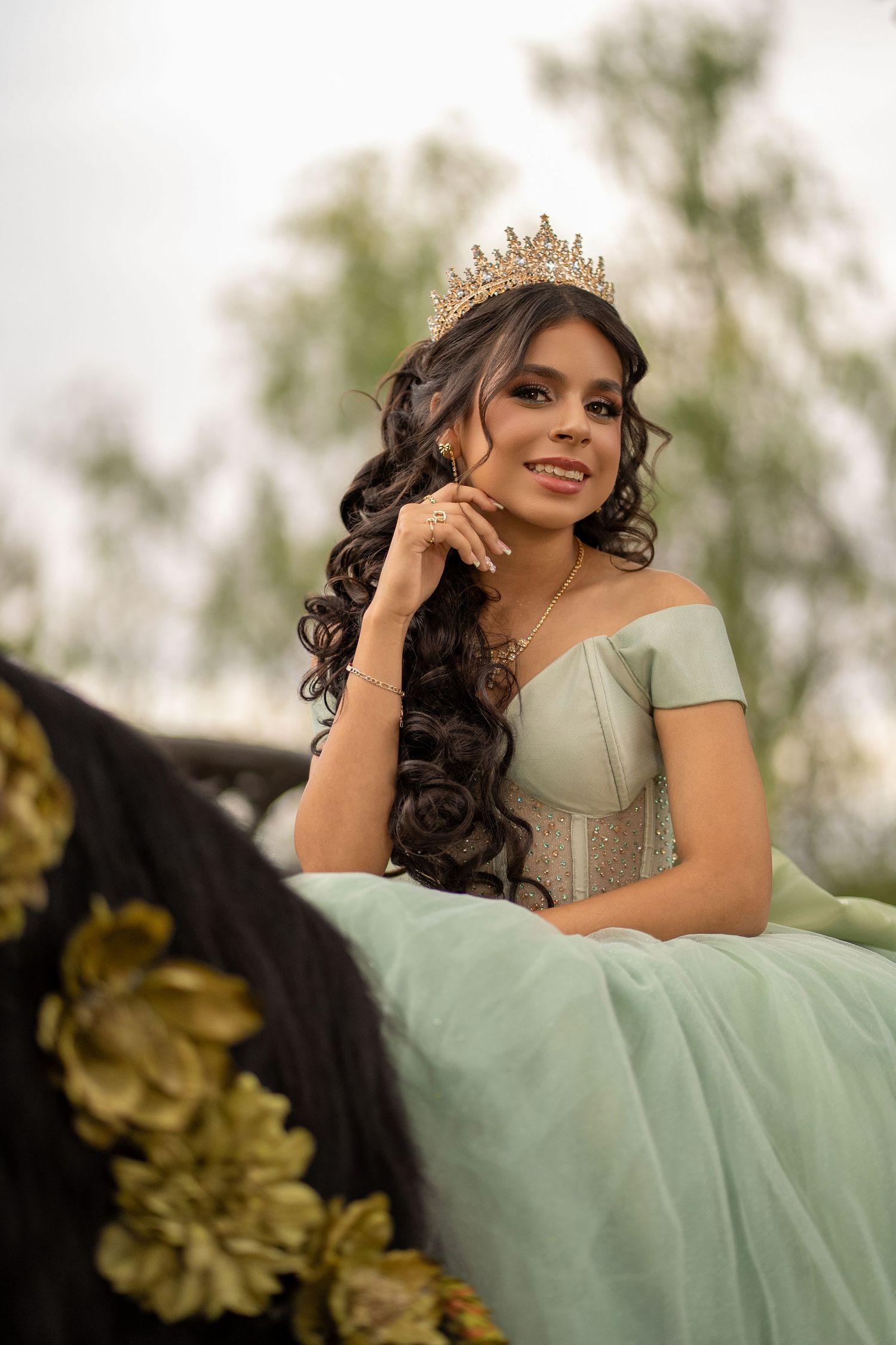 Young woman in mint green quinceañera gown and gold tiara smiles gracefully, posing outdoors near a decorated horse.