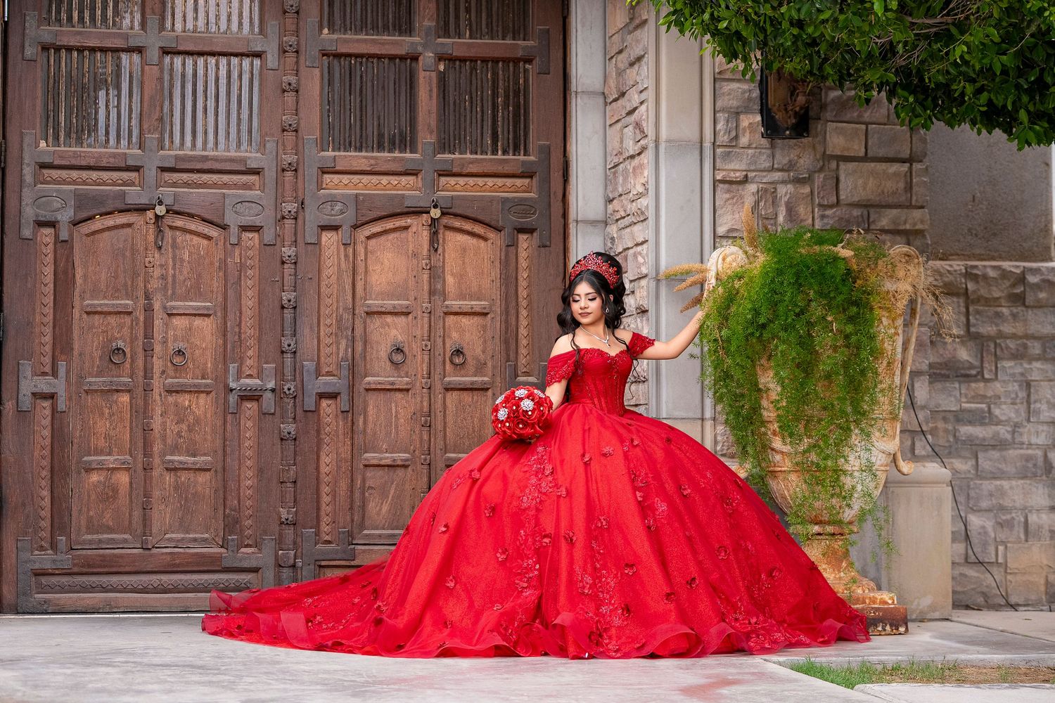 Young woman in red ball gown quinceañera dress poses with bouquet before ornate wooden doors.