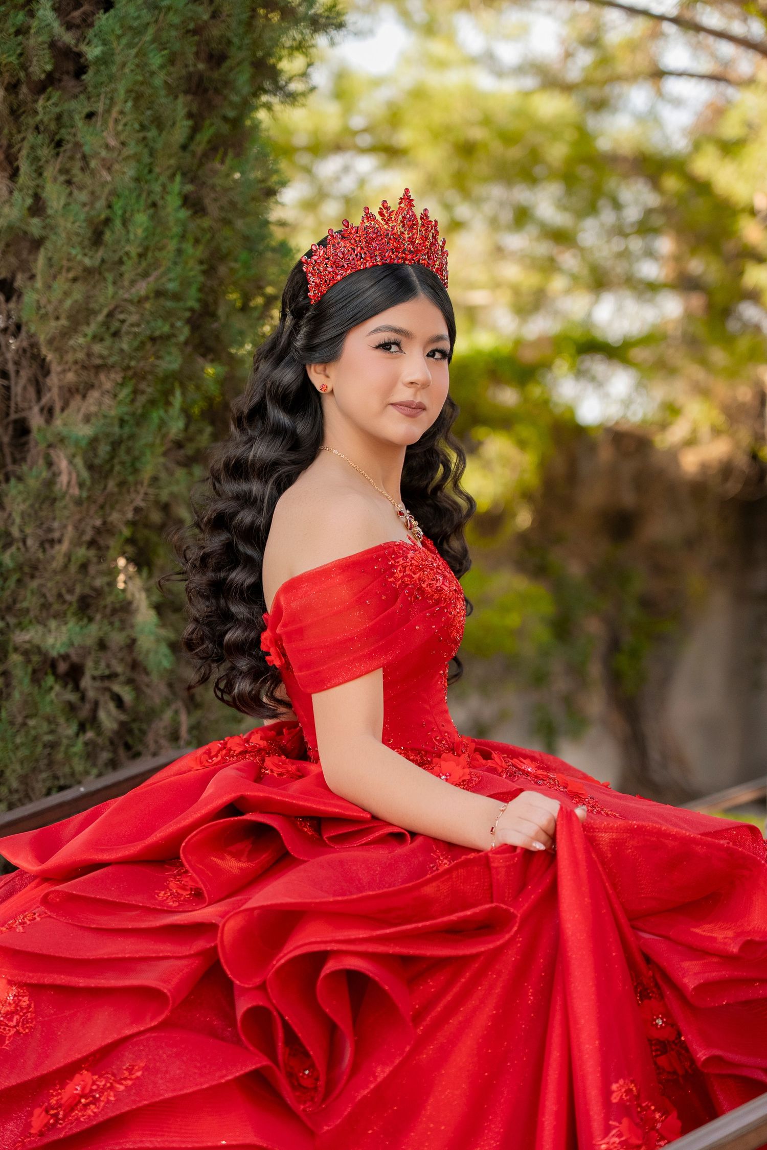 Young woman in red off-shoulder quinceañera gown with red crown poses outdoors among green trees.