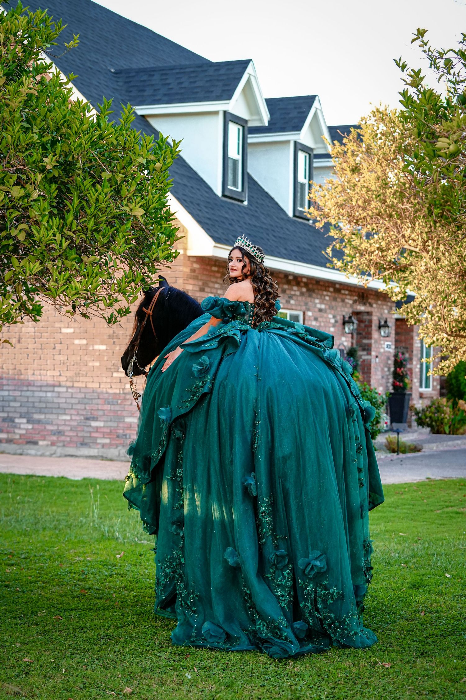 Young woman in an emerald green quinceañera ball gown rides a horse in a residential backyard setting.