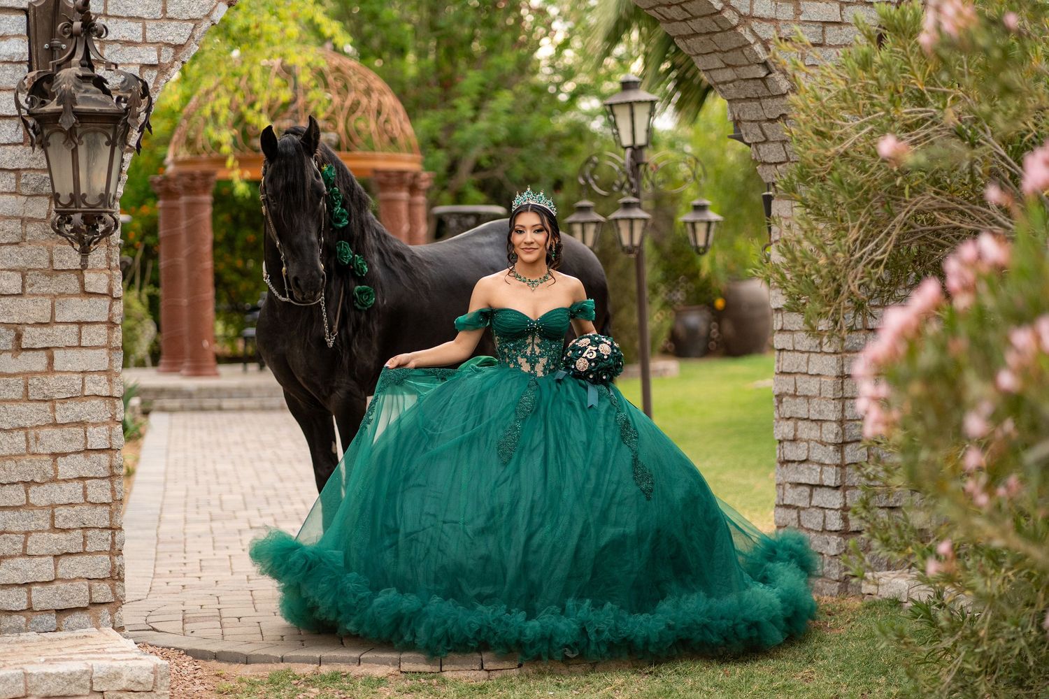 Young woman in emerald green off-shoulder ball gown quinceañera dress poses with black horse under stone archway.