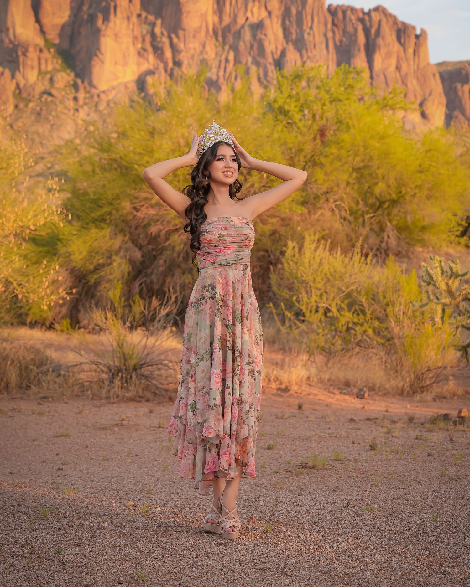 Young woman in floral dress and tiara poses in desert landscape with rocky mountains and green shrubs at golden hour.