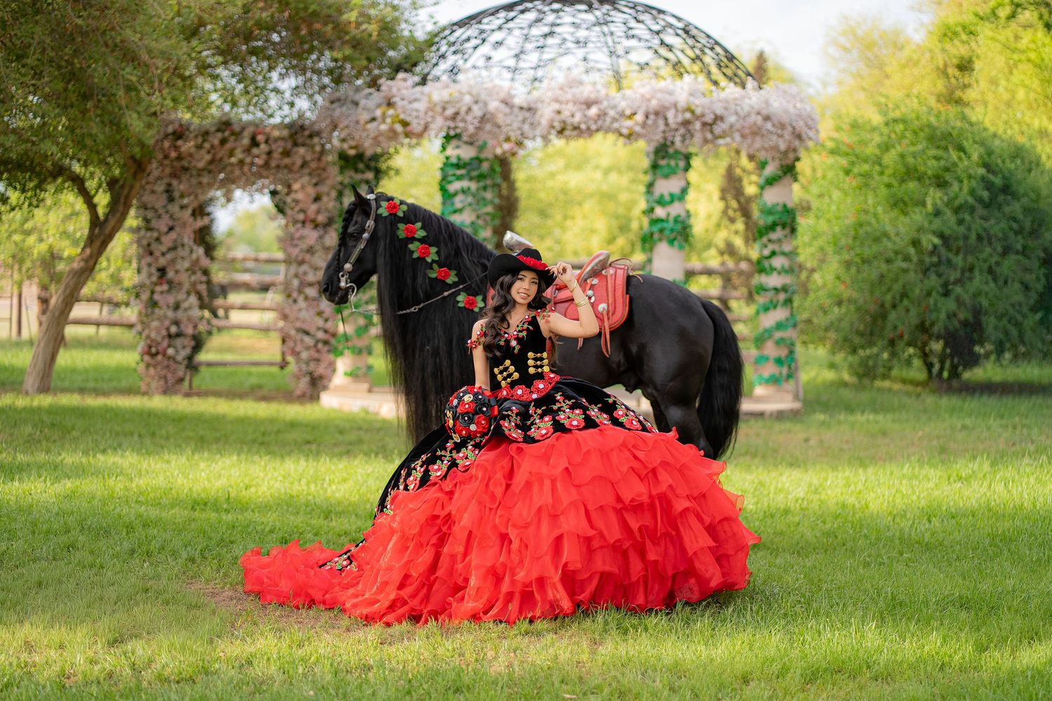 Girl in red quinceañera gown poses with black horse under floral arch in lush green park setting.