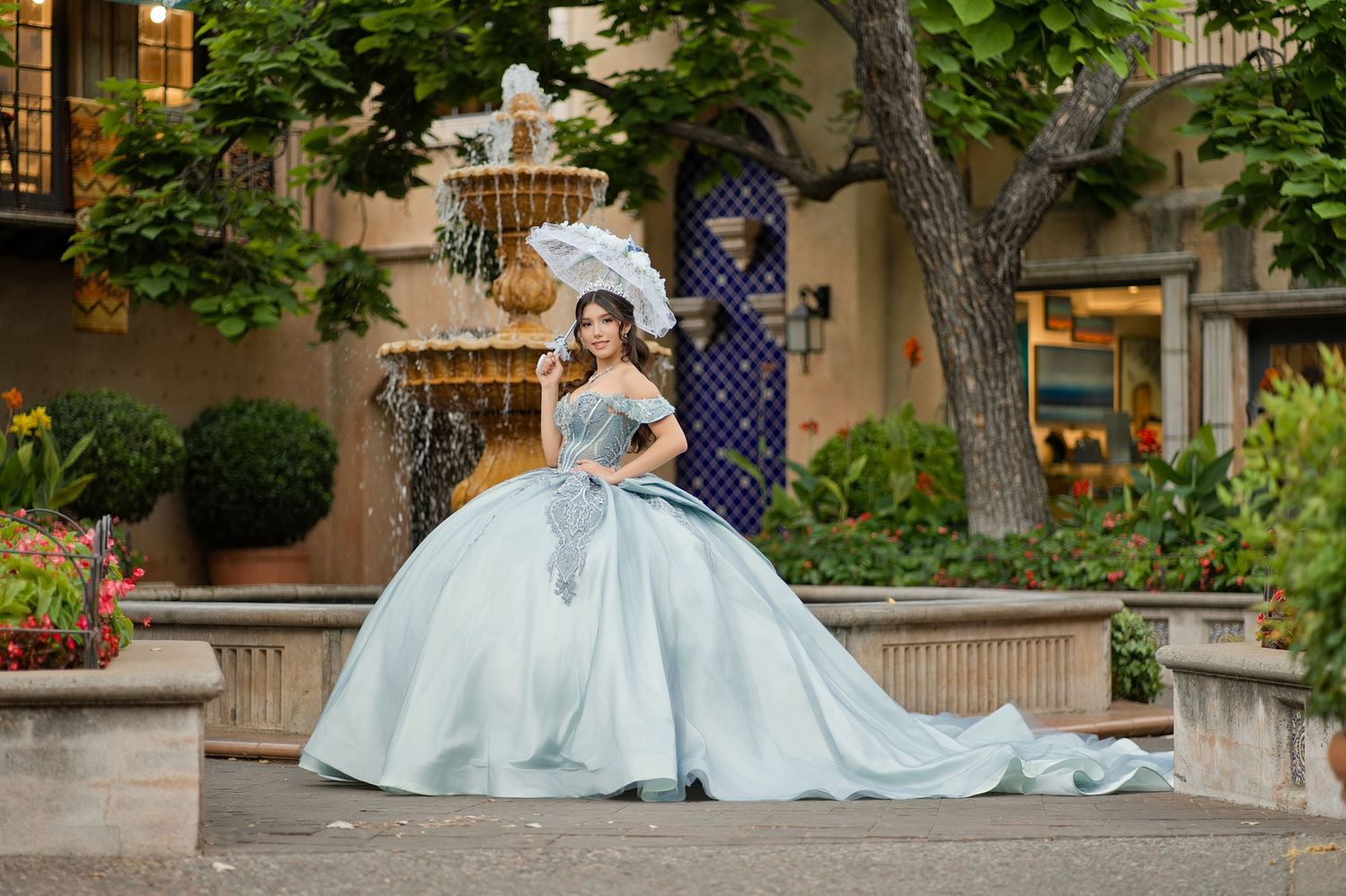 Young woman in elegant light blue quinceañera ball gown with white parasol poses by fountain.