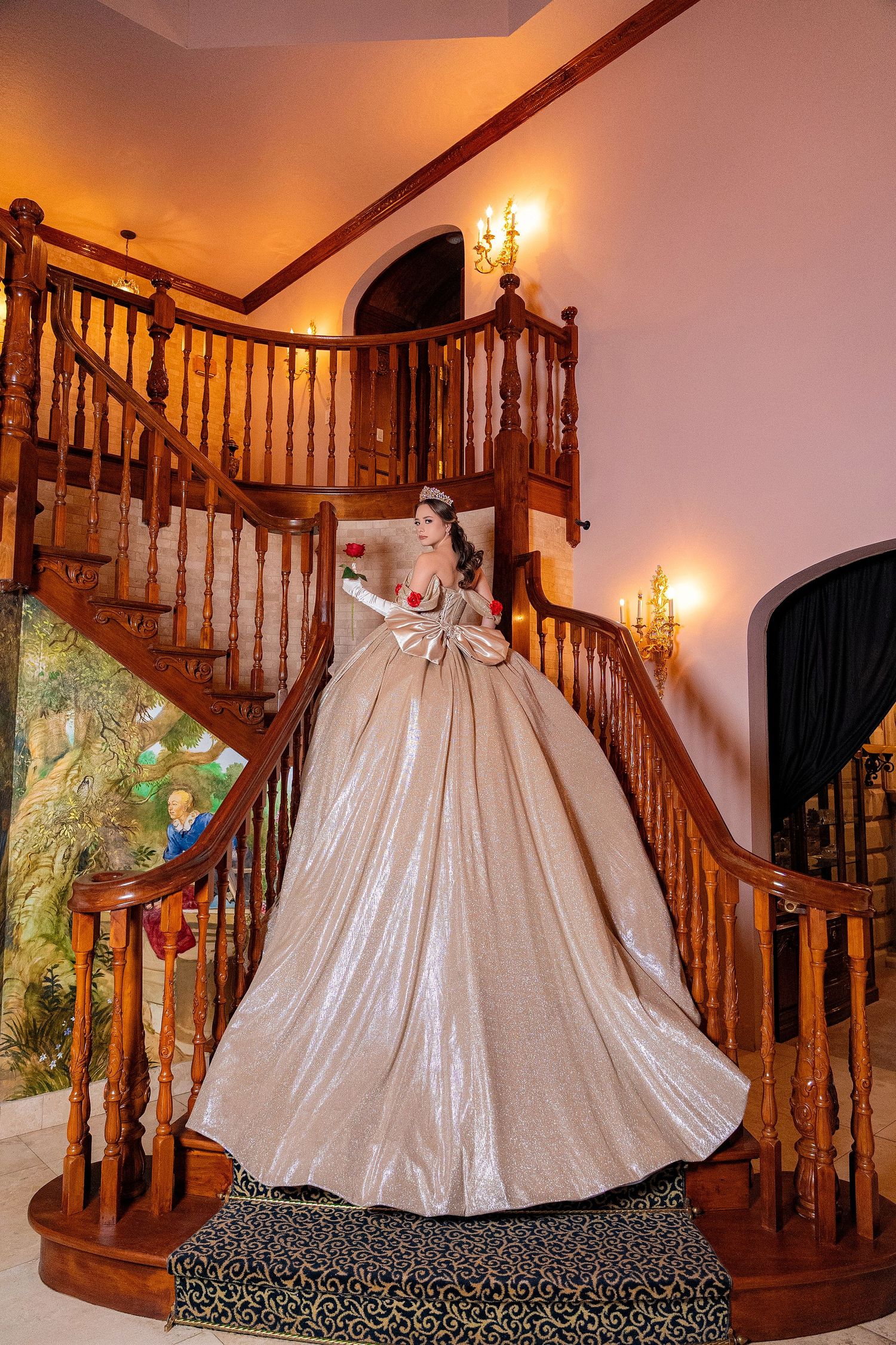 Woman in a stunning golden ball gown stands on an elegant wooden spiral staircase in a grand interior.