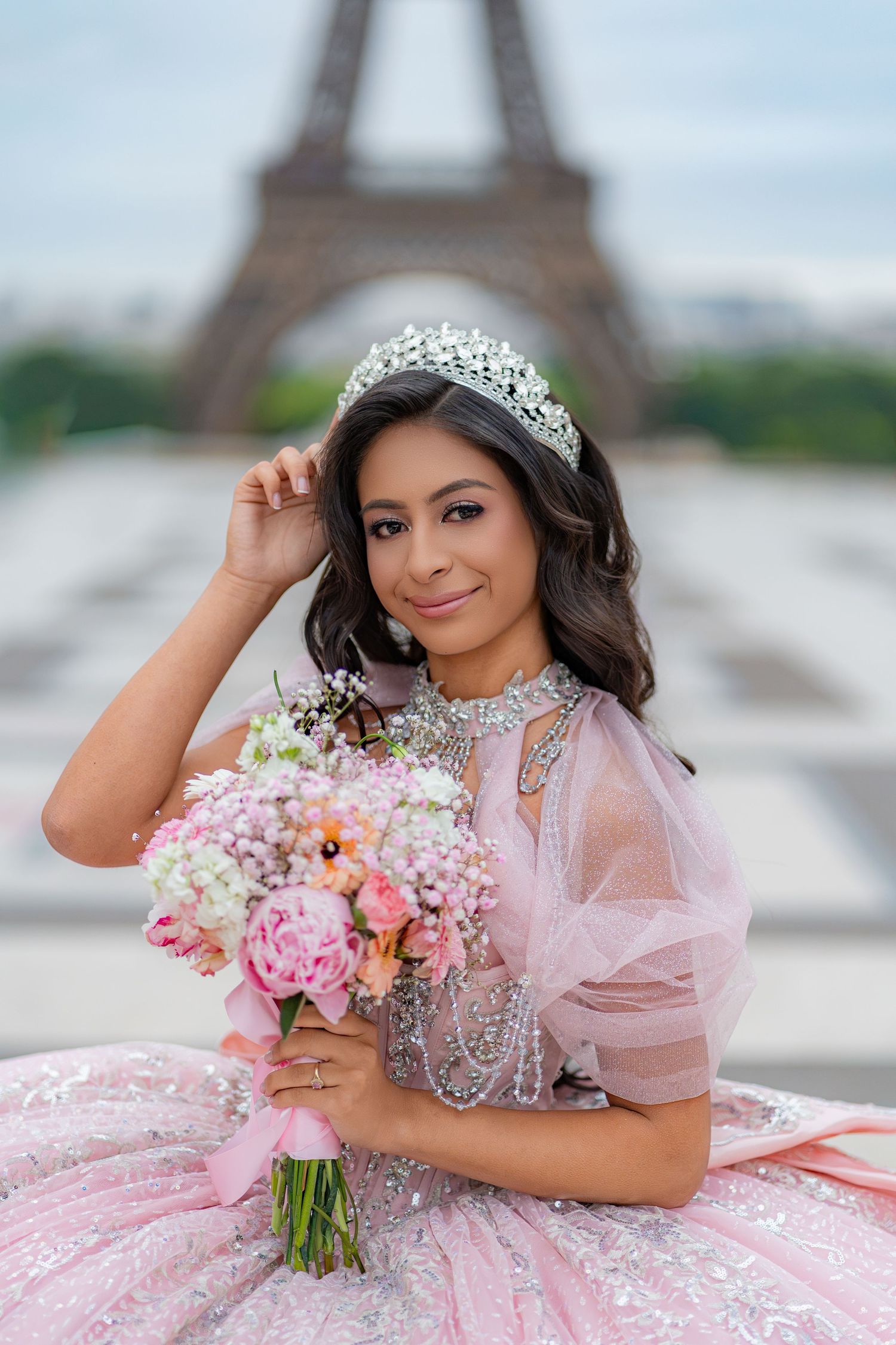 Smiling young woman in pink quinceañera gown holds floral bouquet before the Eiffel Tower.
