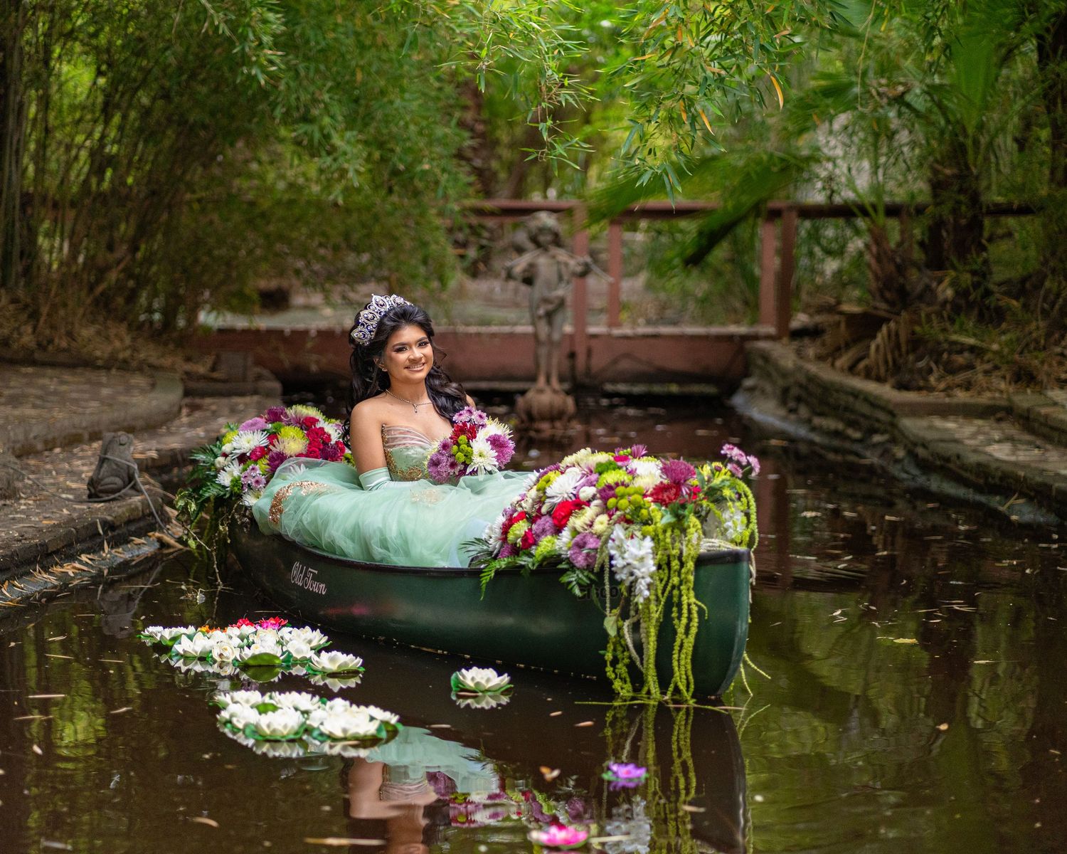 Quinceañera in a mint green gown sits in a flower-adorned canoe surrounded by water lilies in a pond.