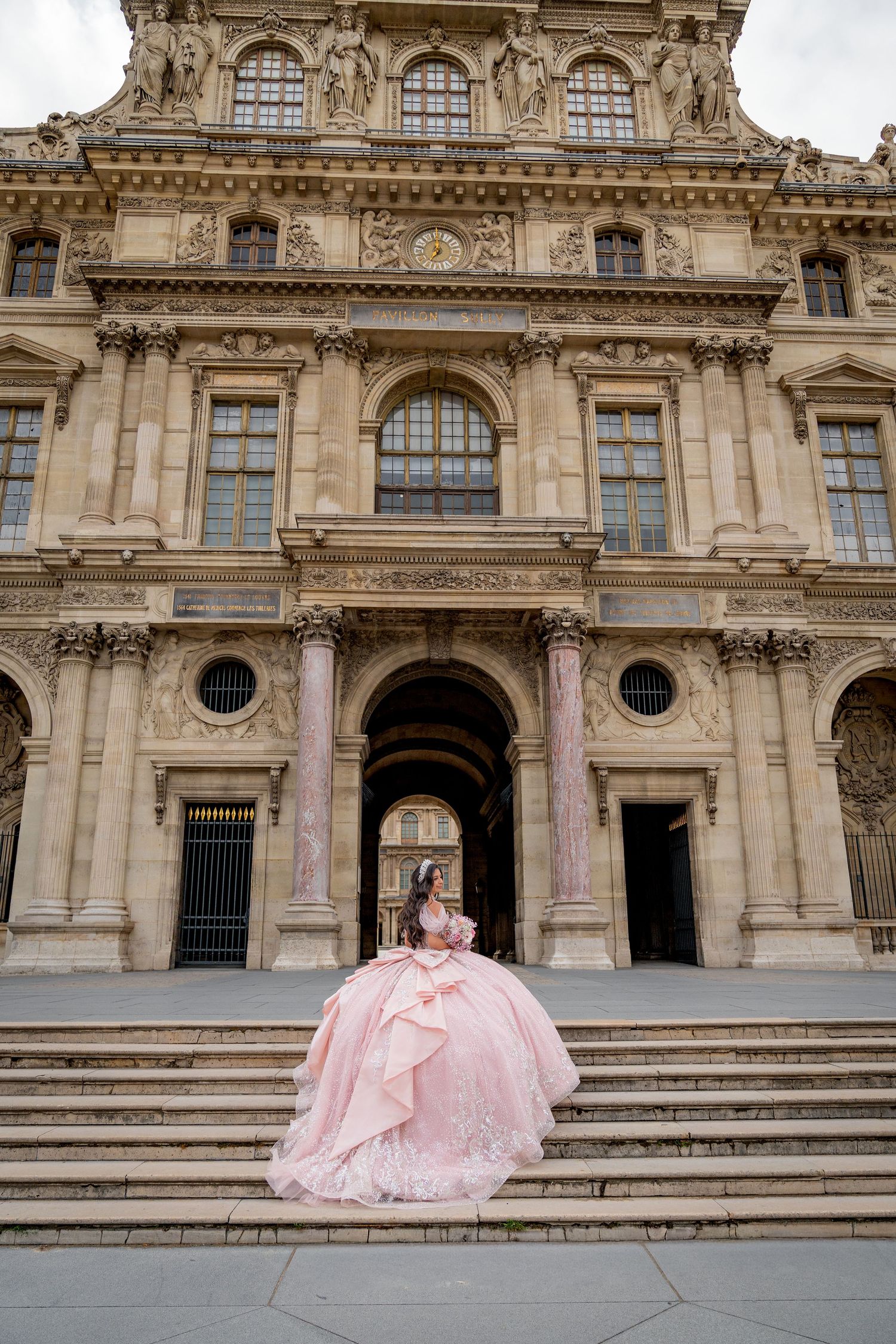Elegant woman in a voluminous pink ball gown poses on steps outside the grand Louvre Museum in Paris.