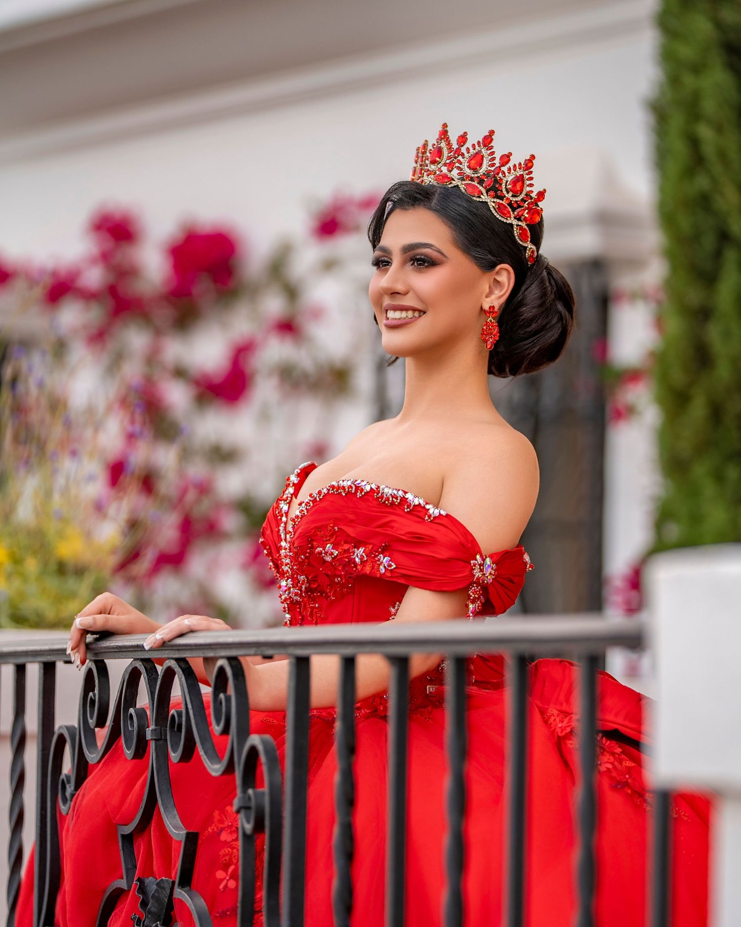 Smiling young woman in red off-shoulder quinceañera gown and crown poses at iron garden railing.