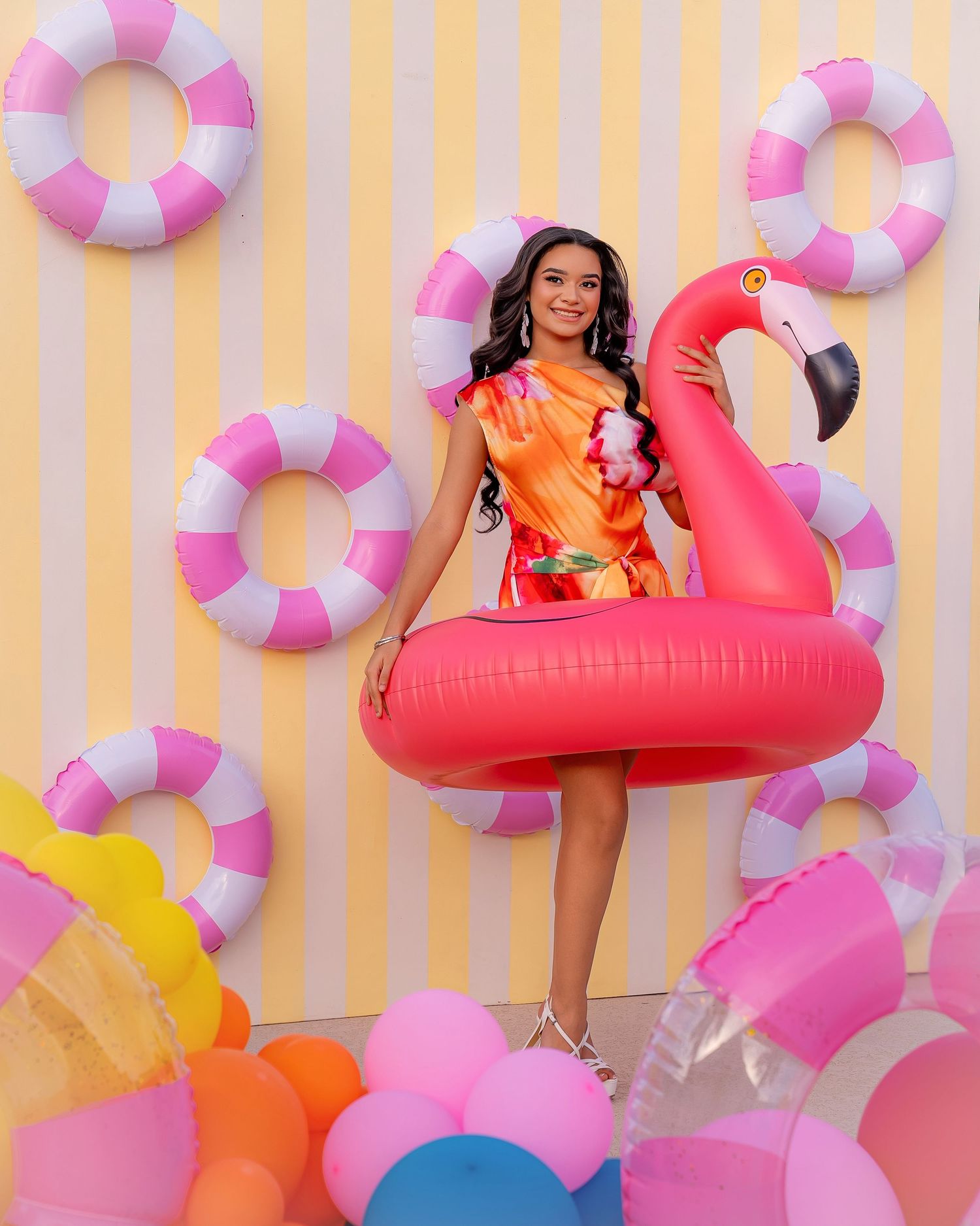 Woman in orange dress holding pink flamingo pool float surrounded by colorful balloons and life rings on yellow striped backdrop.