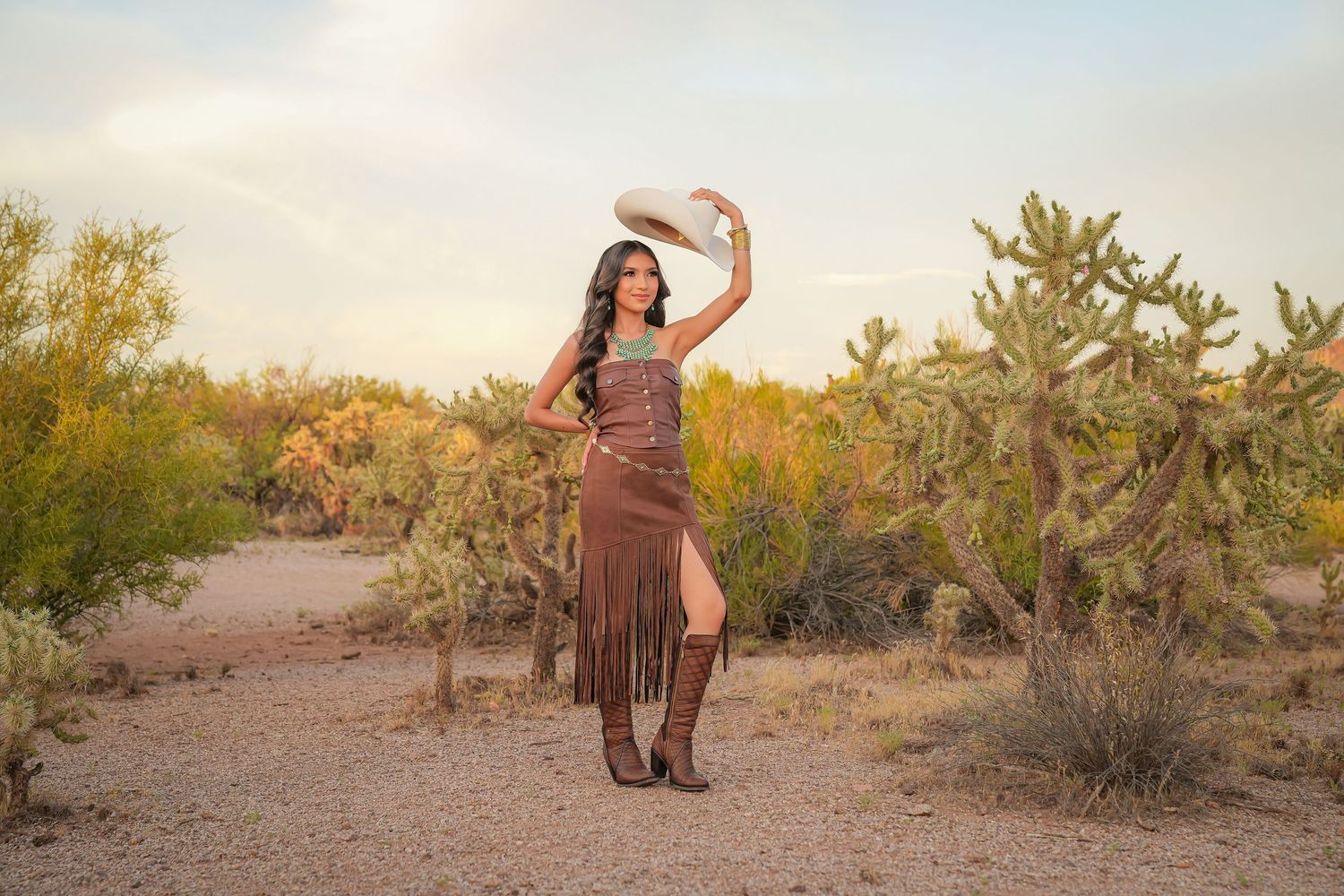 Woman in brown fringe dress and cowboy hat poses among cacti in desert landscape at sunset.