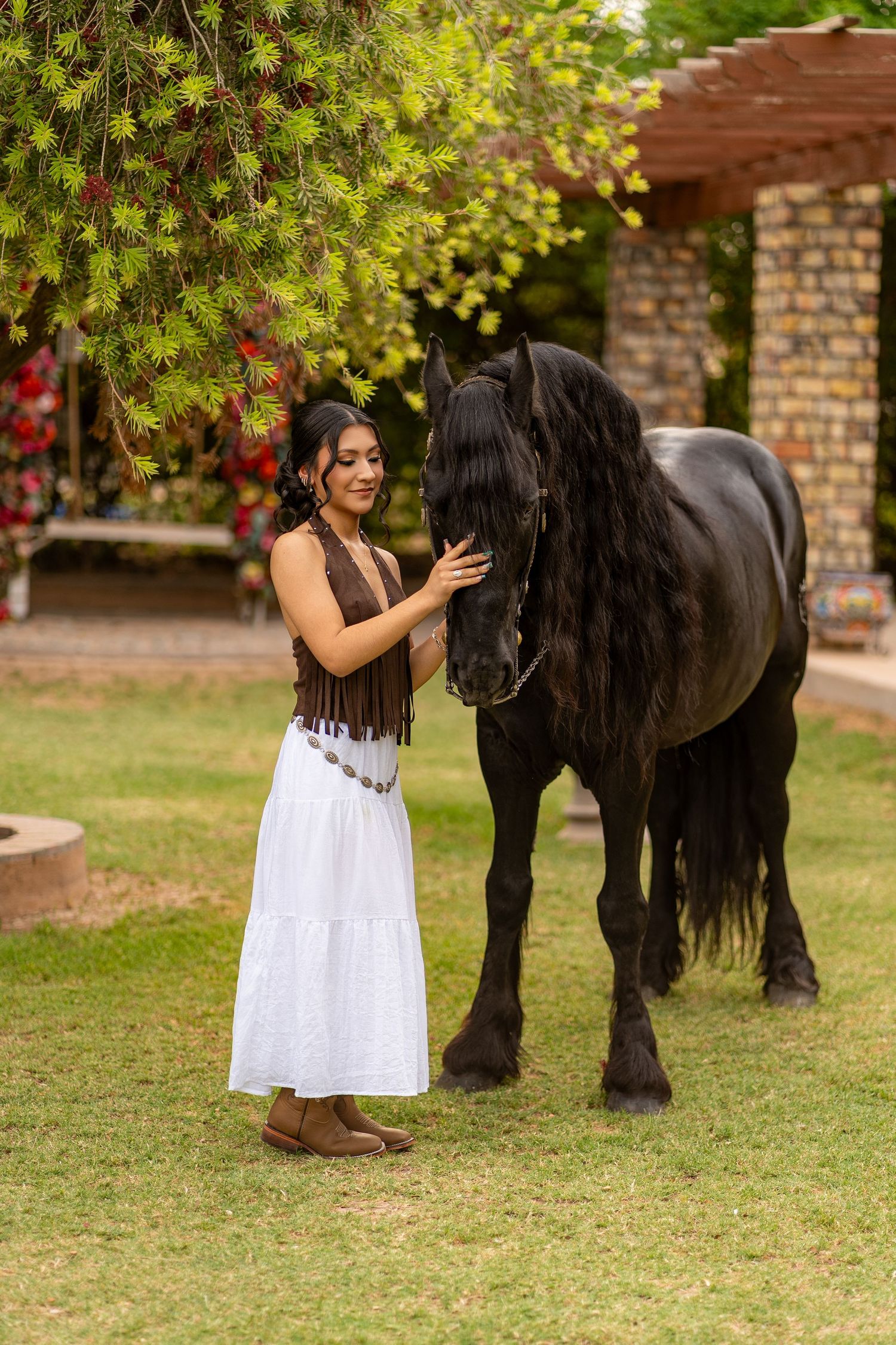 A woman in a white skirt and brown fringe top gently pets a black Friesian horse on a green lawn.