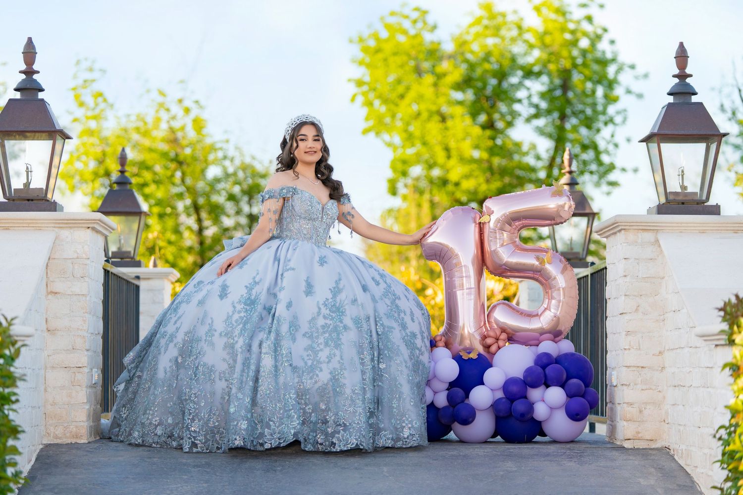 Young woman in blue ballgown poses with balloon number 15 for her quinceañera celebration outdoors.