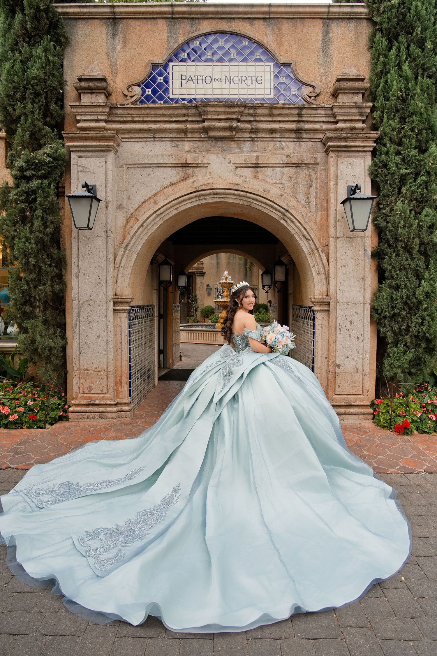 Young woman in a stunning light blue ball gown with long train poses before ornate stone arch at Patio del Norte.