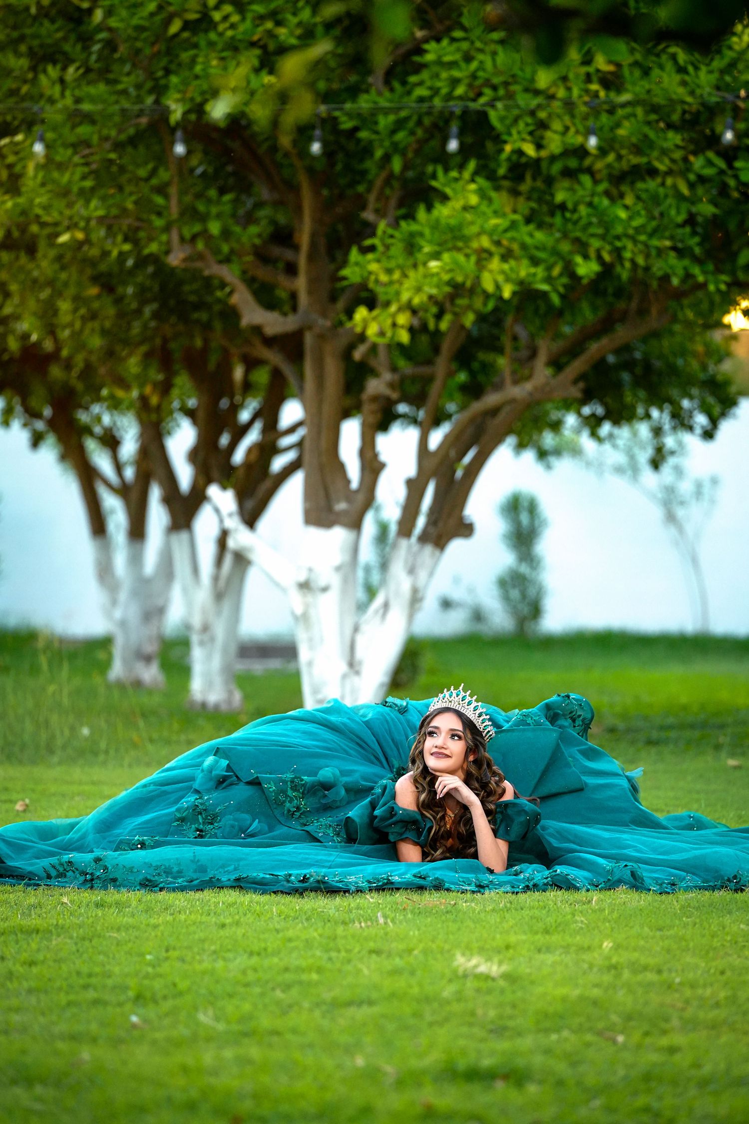 Young woman in a teal quinceañera ball gown with tiara lies gracefully on green grass beneath a tree.