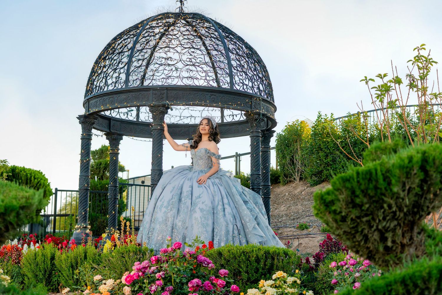 Young woman in elegant blue ball gown poses under ornate gazebo surrounded by colorful garden flowers.