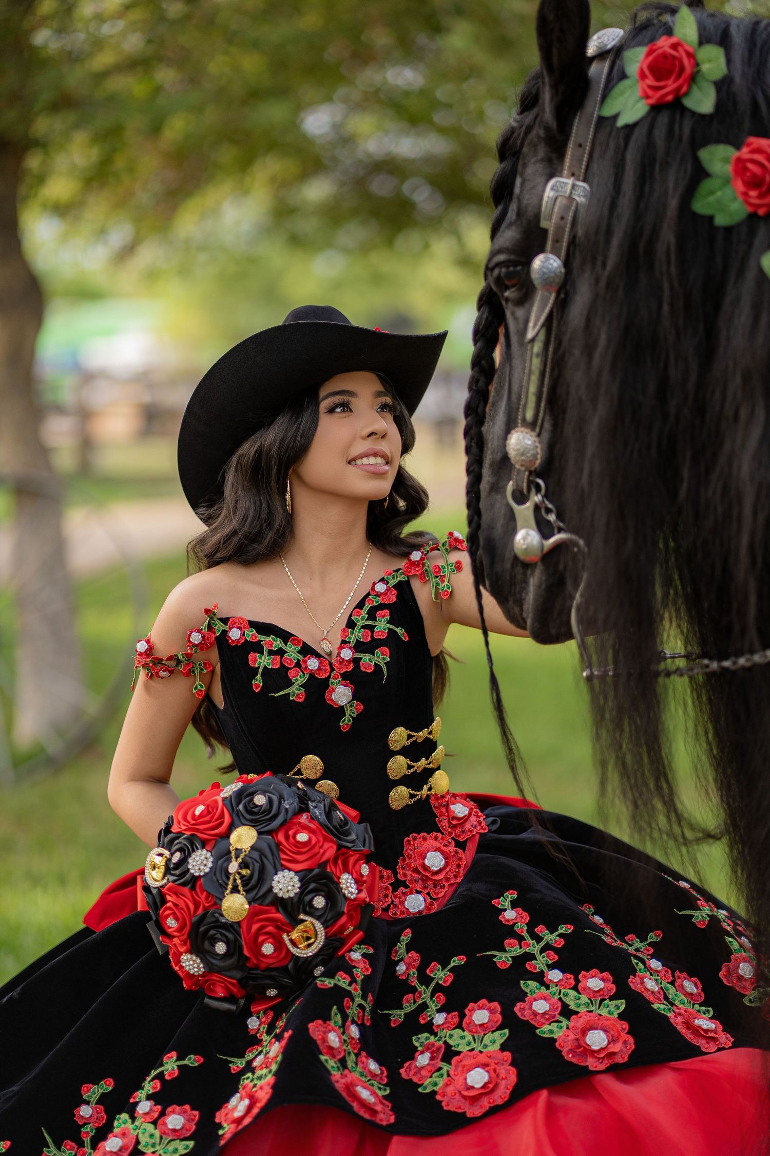 Young woman in black floral embroidered quinceañera gown and cowboy hat poses beside a decorated black horse outdoors.