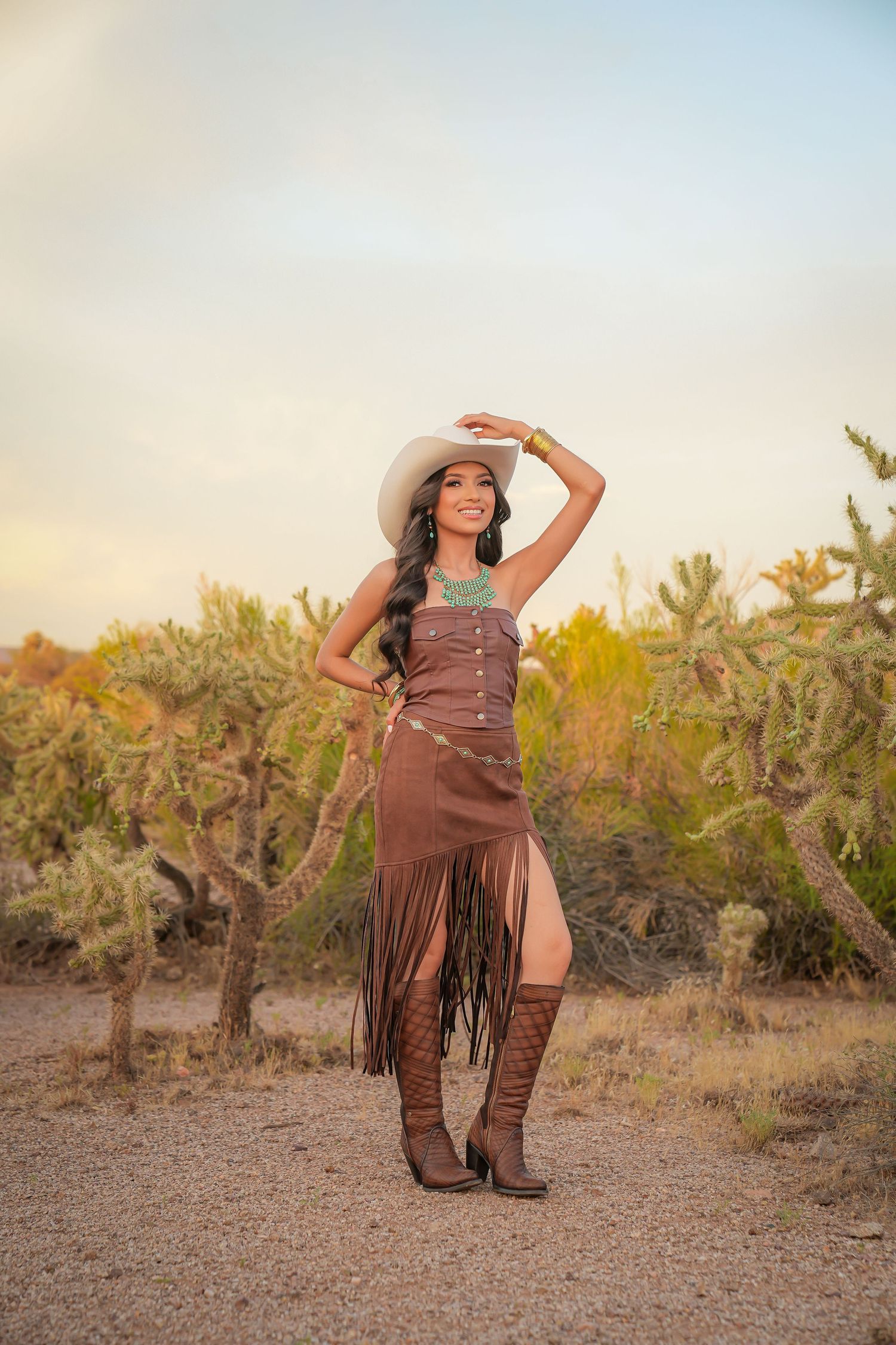 Woman in cowgirl outfit with white hat, brown fringe dress, and boots poses among desert cacti at golden hour.