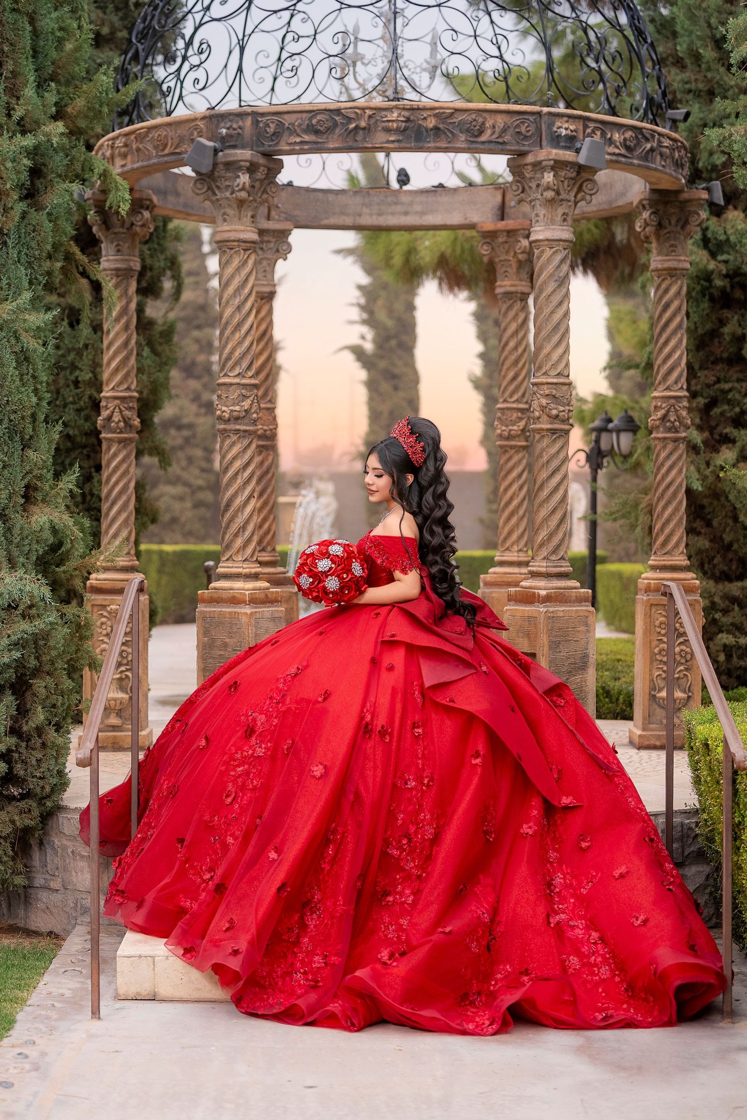 Young woman in a red floral ball gown stands under a stone gazebo in a garden at sunset for her quinceañera.