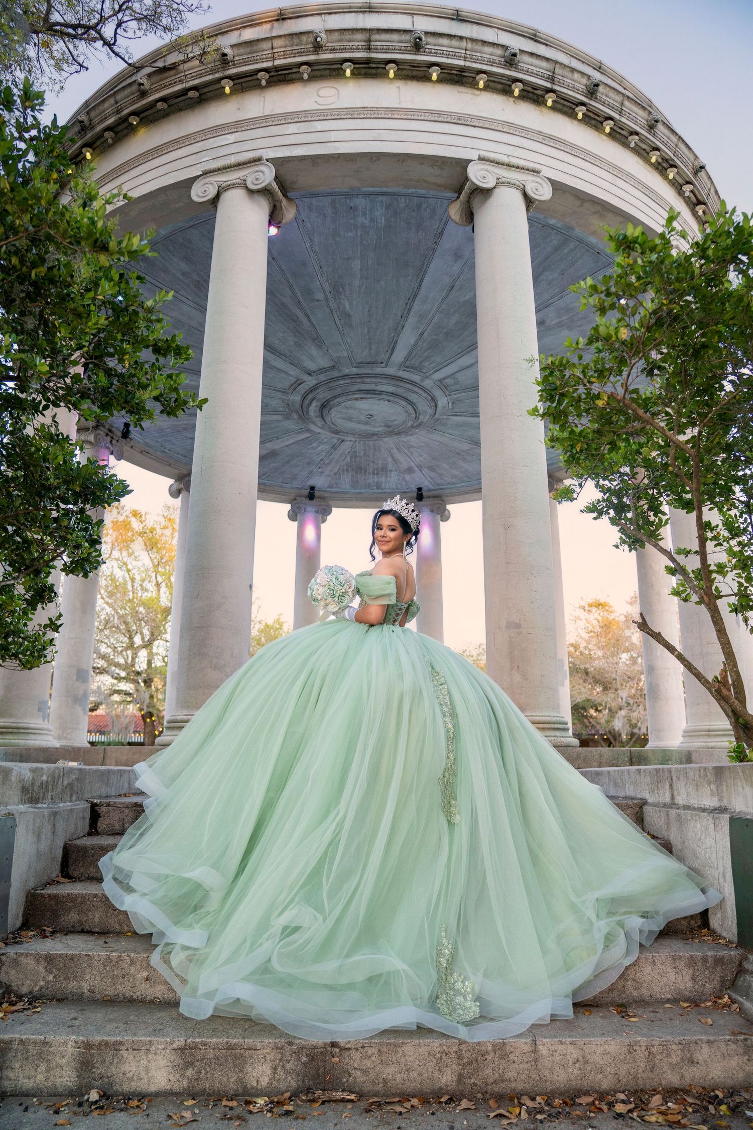 Young woman in a mint green quinceañera ball gown poses on stone steps before a classical rotunda.