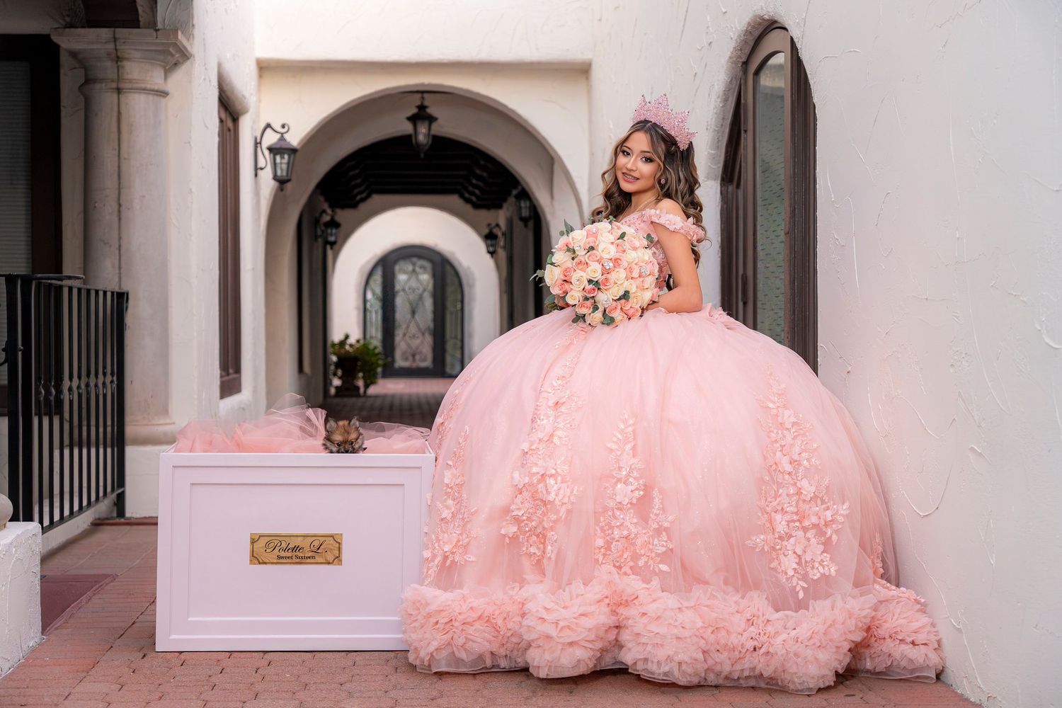Young woman in a pink ball gown quinceañera dress holding a bouquet, posing outdoors near an elegant white building.