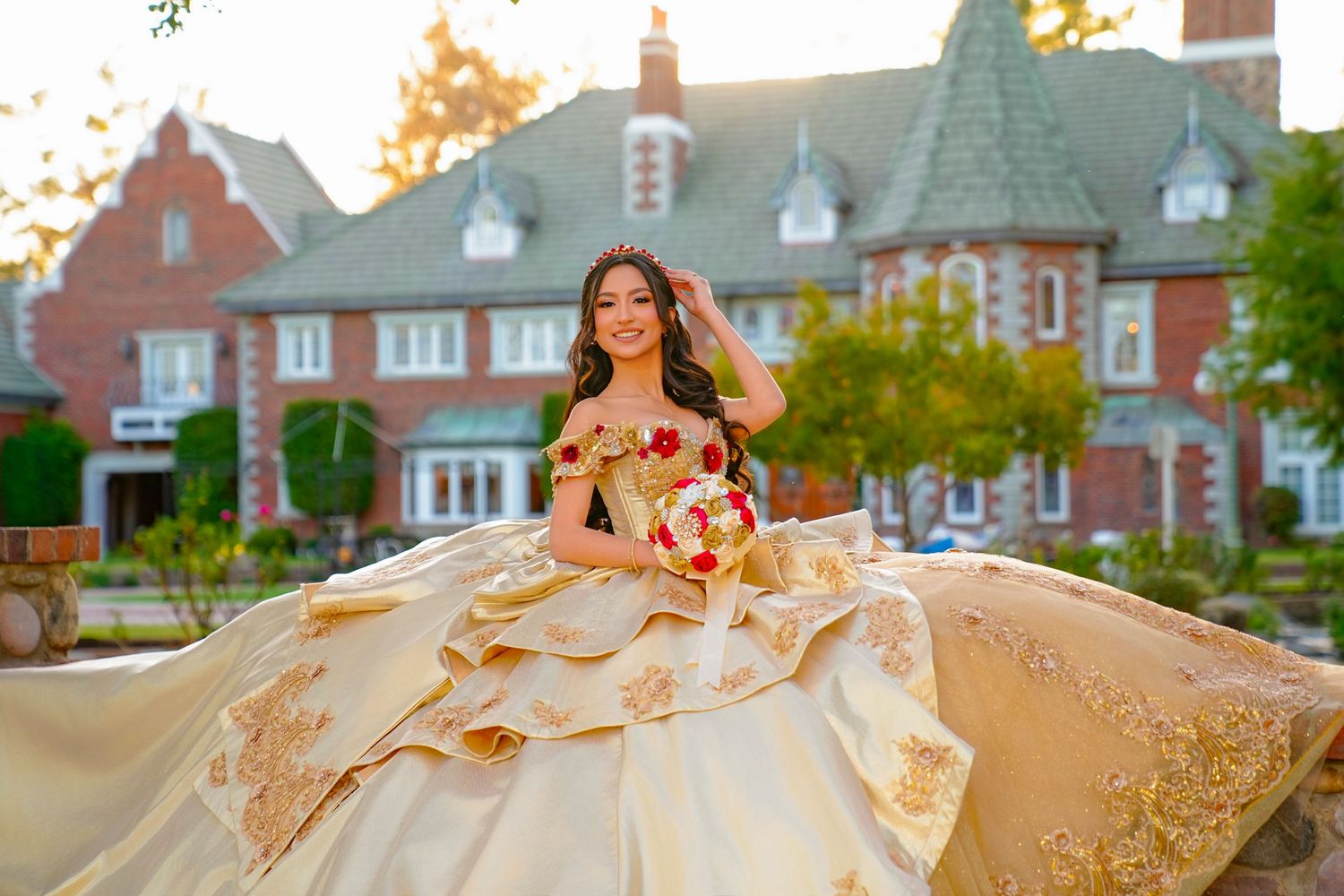 Young woman in a flowing gold quinceañera ball gown twirling before a grand brick estate at sunset.