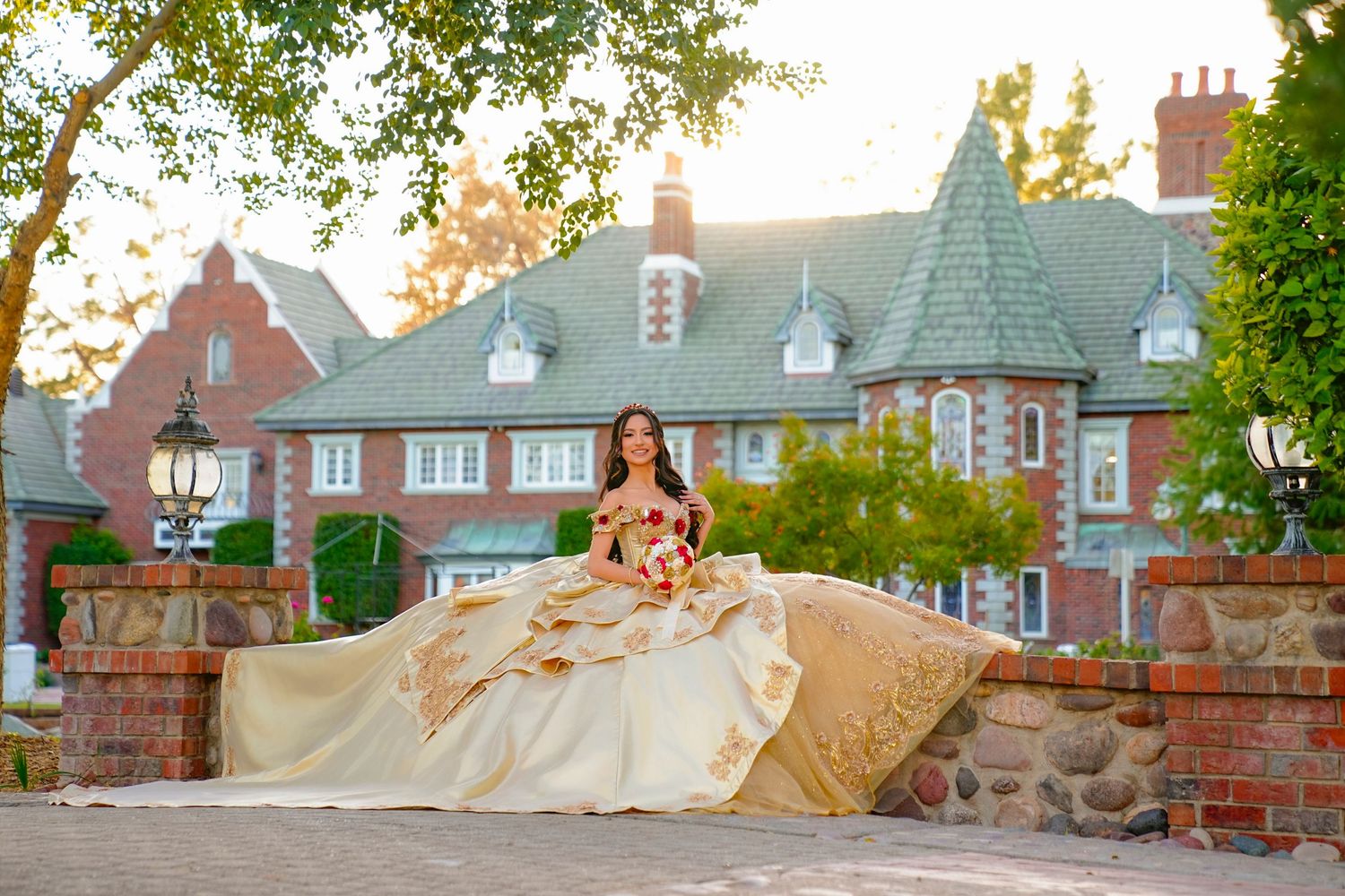 Young woman in elegant gold ball gown poses on stone wall before a grand brick mansion at sunset.
