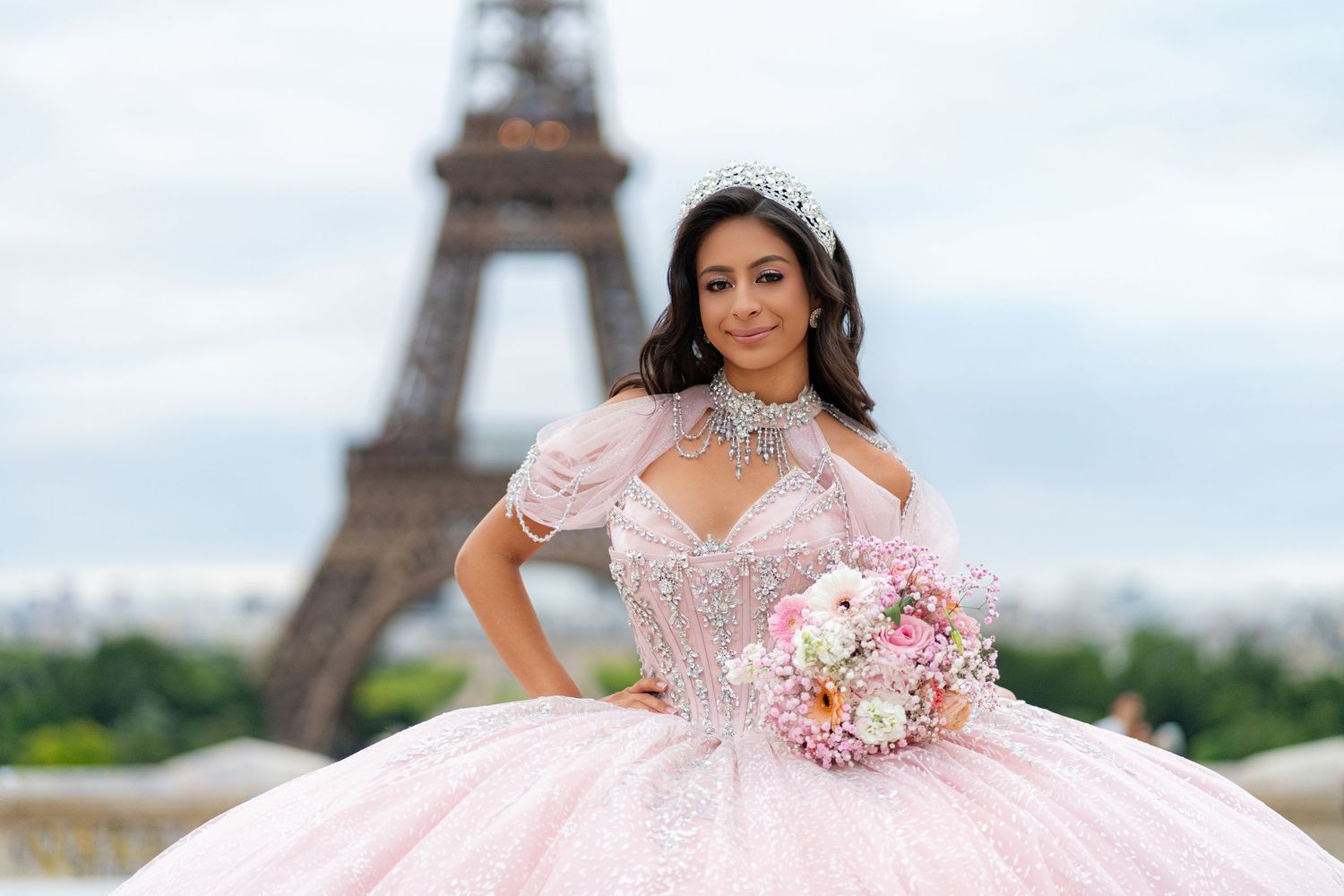 Young woman in pink quinceañera gown with tiara holds bouquet before the Eiffel Tower in Paris.