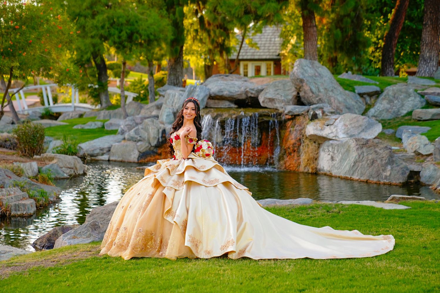 Elegant young woman in a layered gold quinceañera gown standing near a rocky waterfall garden pond.
