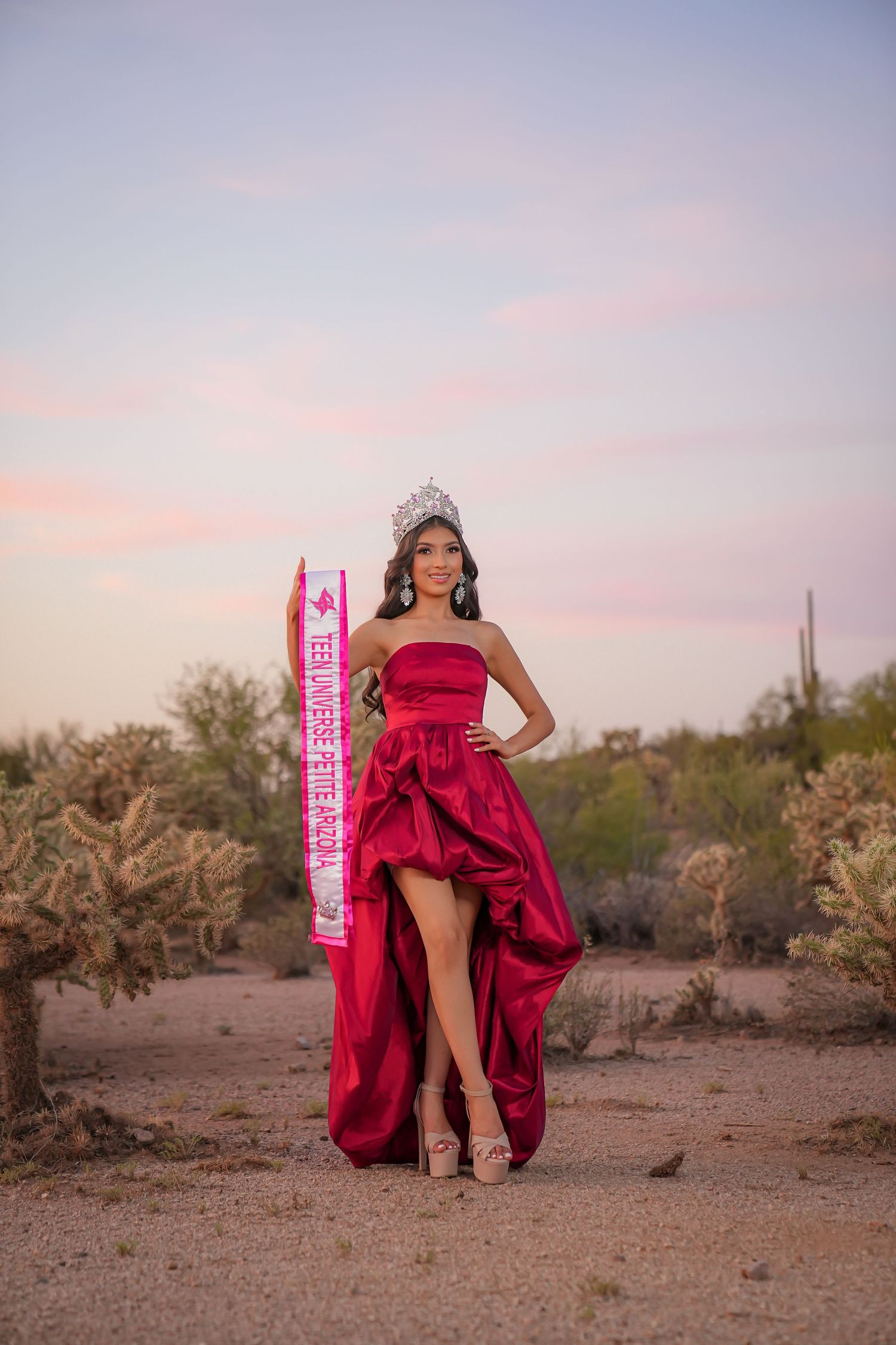 Young woman in red gown wearing crown and sash poses in desert landscape at sunset.
