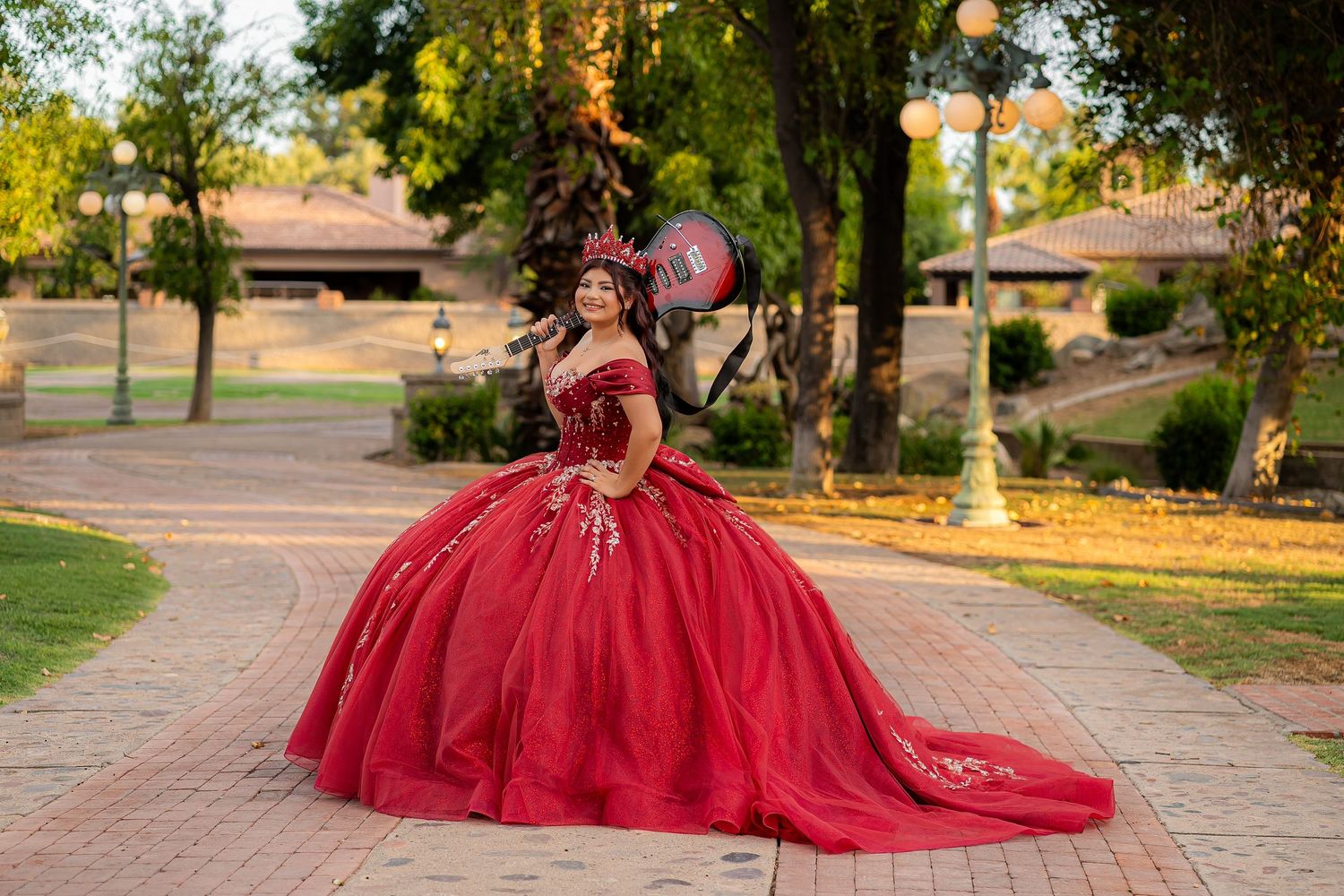 Young woman in a stunning red ballgown quinceañera dress poses on a brick path surrounded by lush green trees.