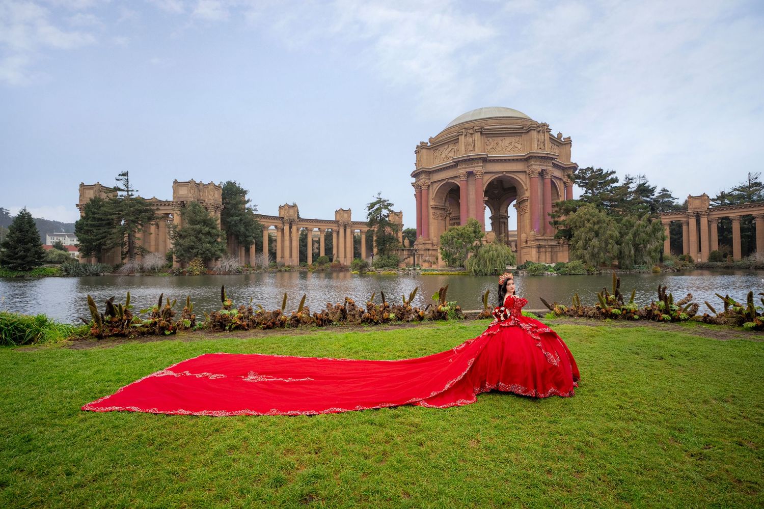 Woman in red ballgown with long train poses on grass at Palace of Fine Arts, San Francisco.