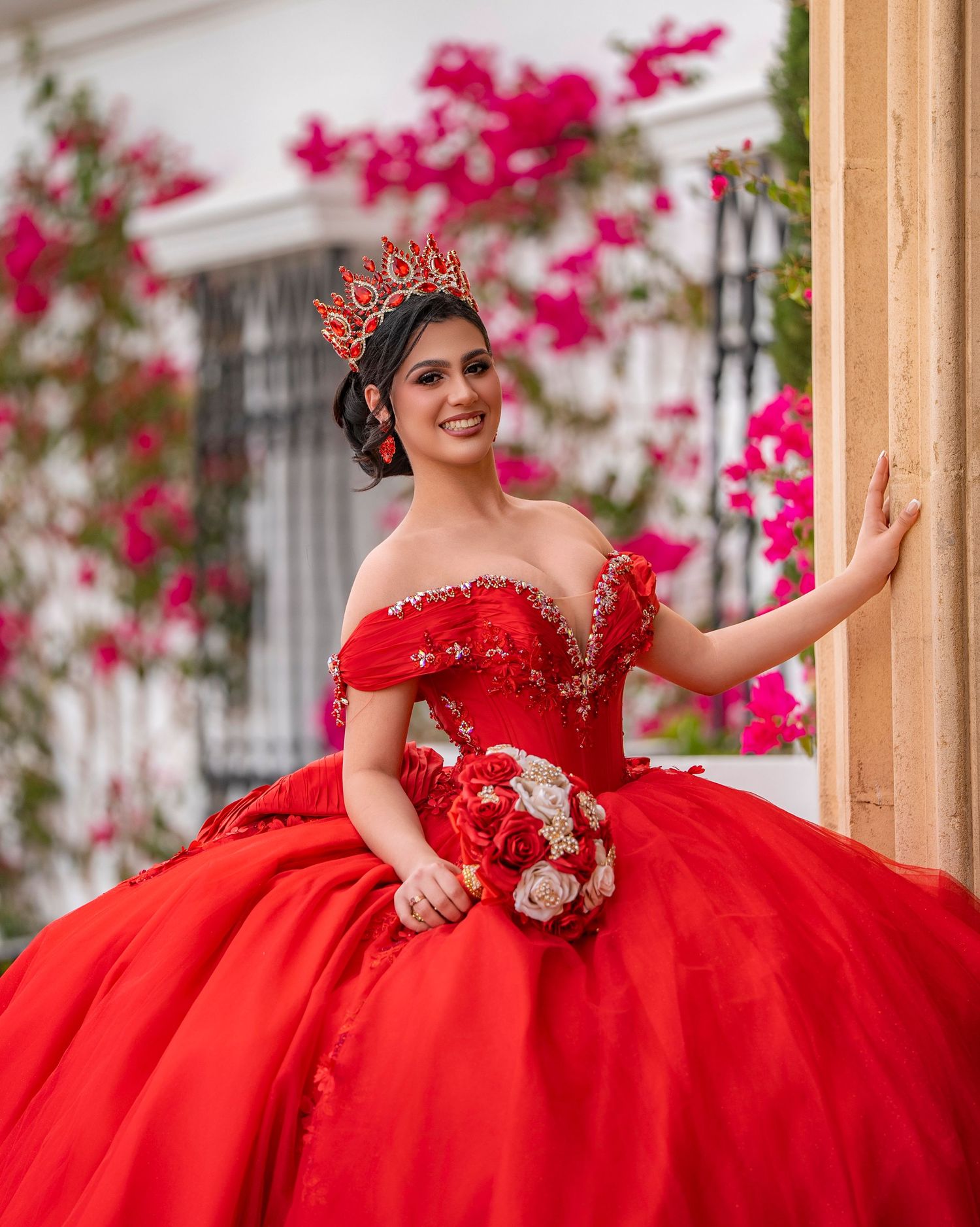 Young woman in a red ball gown with crown and bouquet poses at a quinceañera celebration outdoors.