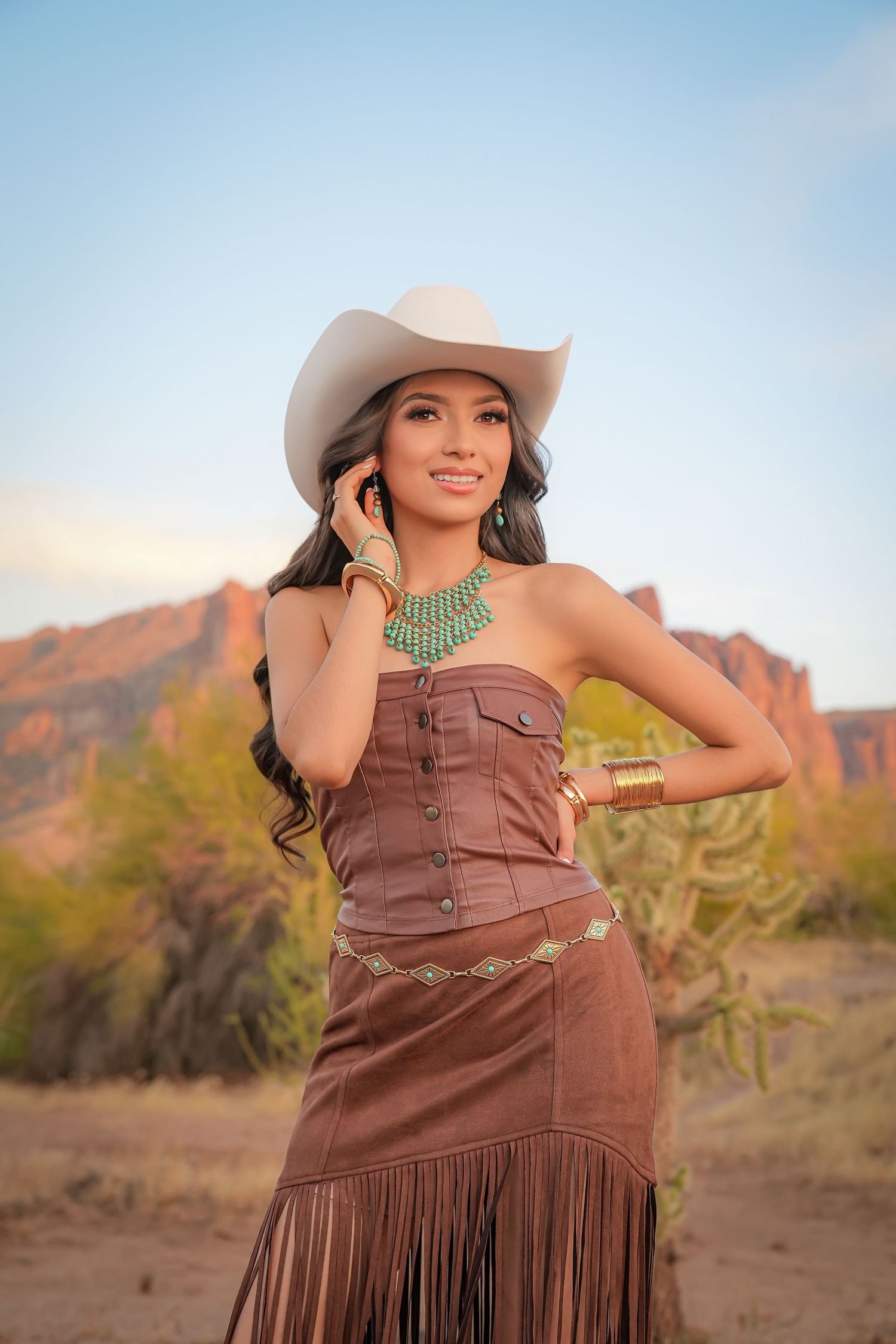 Young woman in cowboy hat and brown fringe dress with turquoise jewelry poses in desert landscape.