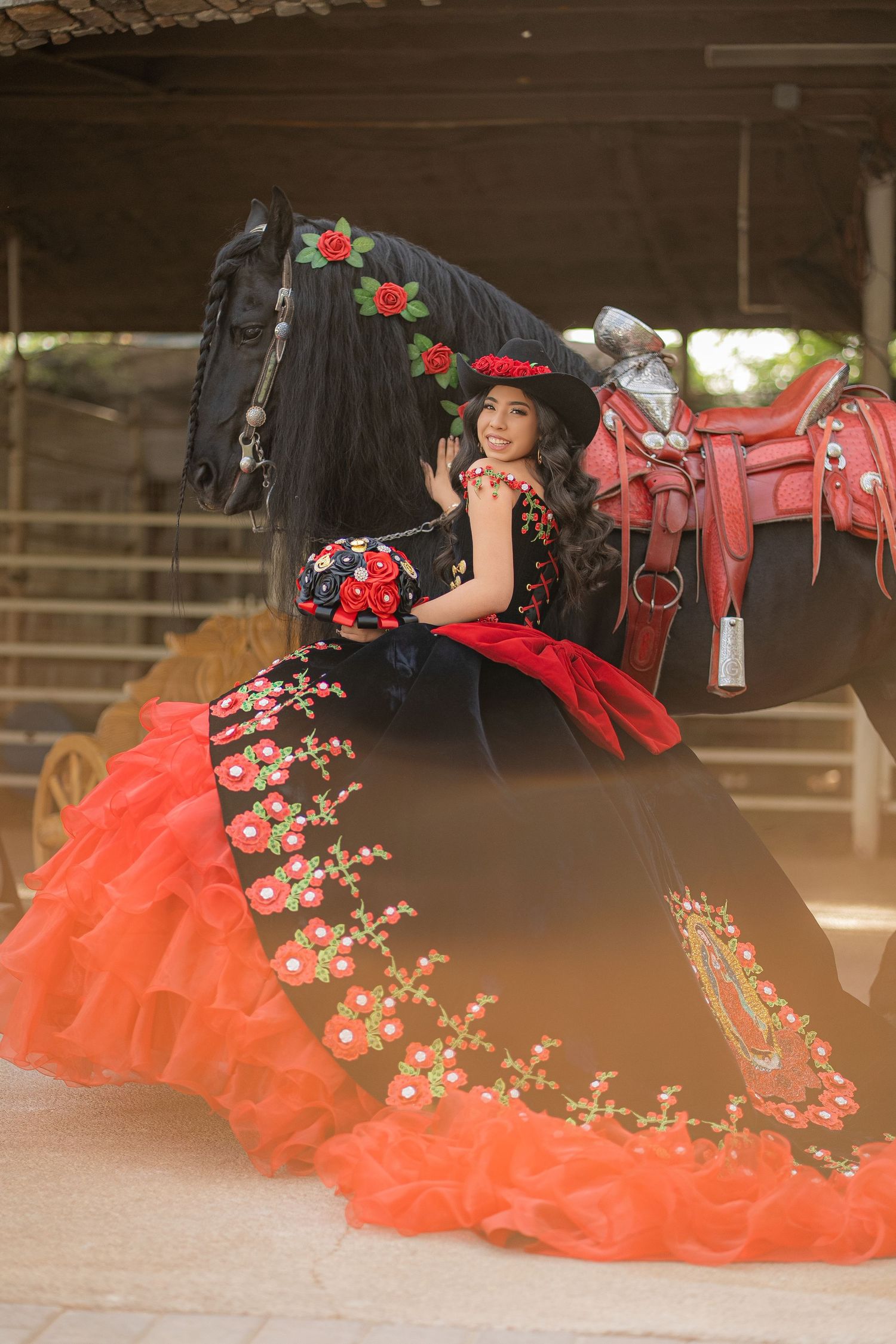 Young woman in red and black floral quinceañera gown poses beside a black horse with red rose decorations.