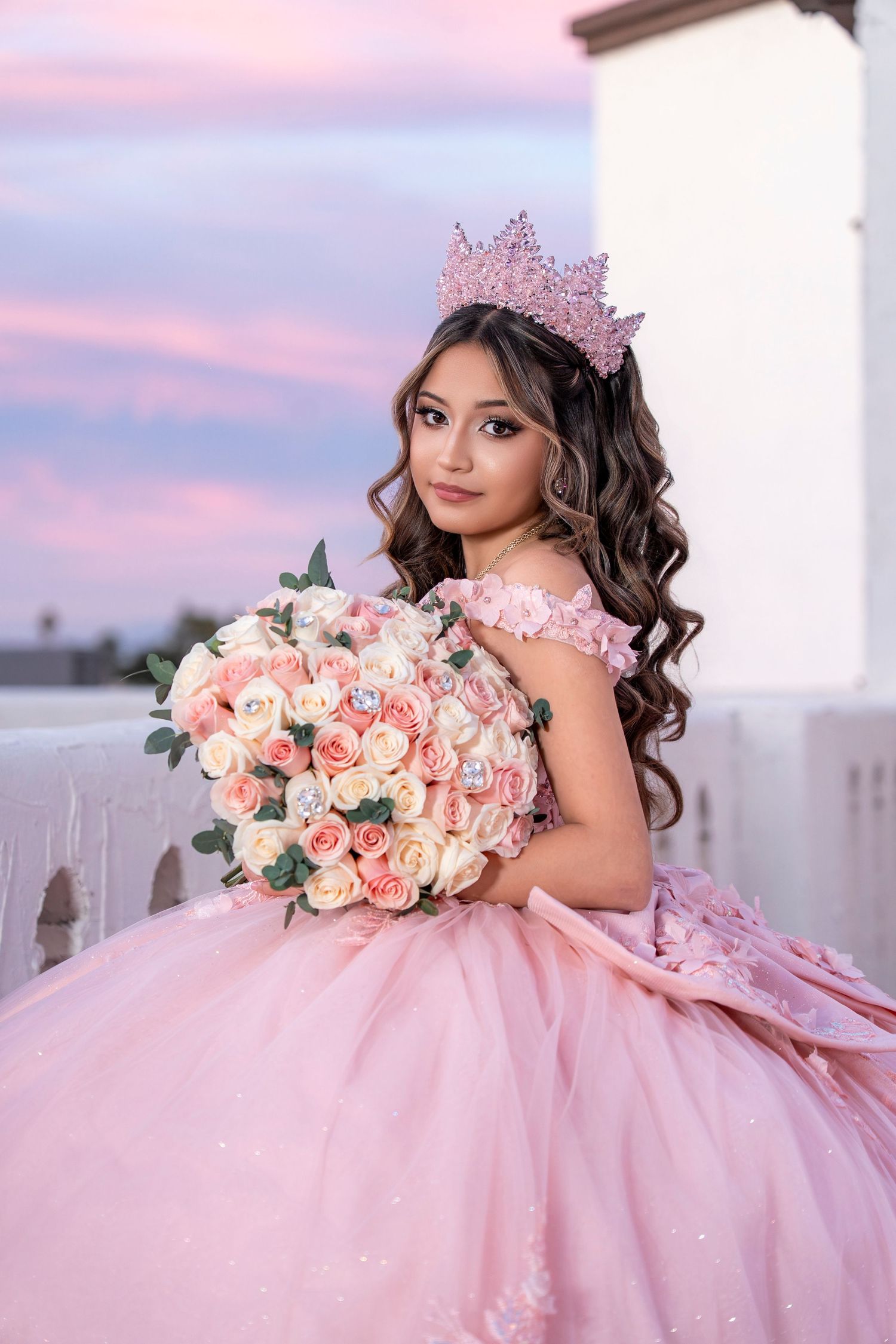 Young girl in pink ball gown and crown holds rose bouquet at sunset quinceañera photo session.