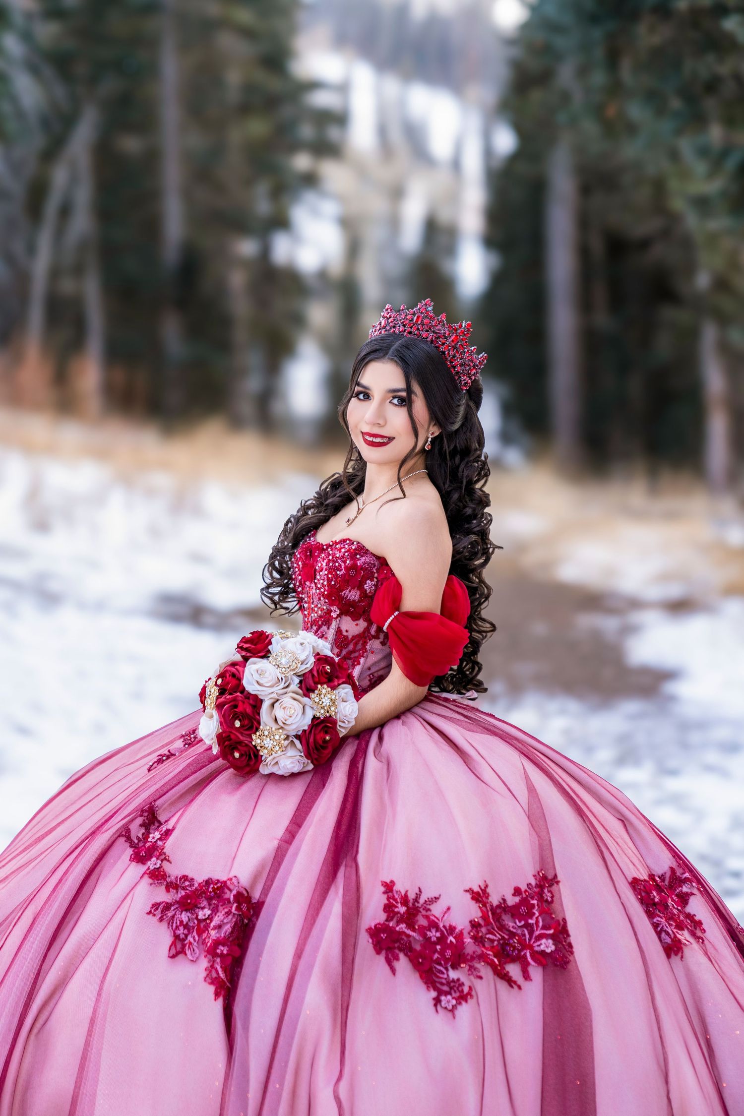 Young woman in a pink and red ball gown with floral appliques holds a bouquet in a snowy winter forest.