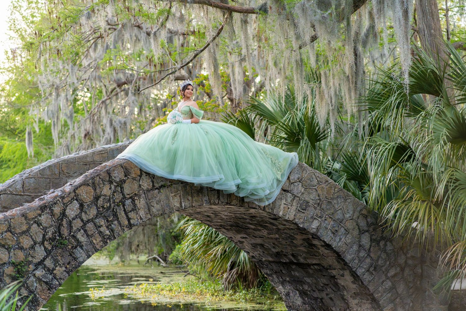Young woman in a flowing mint green quinceañera ball gown poses gracefully on a mossy stone arch bridge.