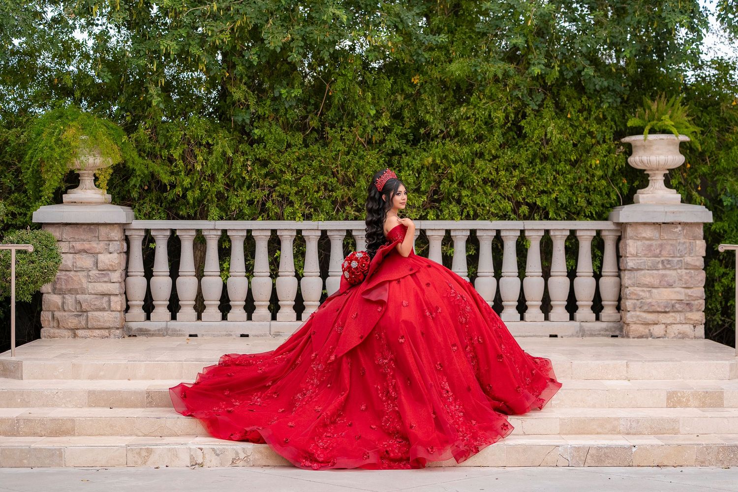 Young woman in a stunning red ballgown quinceañera dress poses on stone steps surrounded by lush greenery.