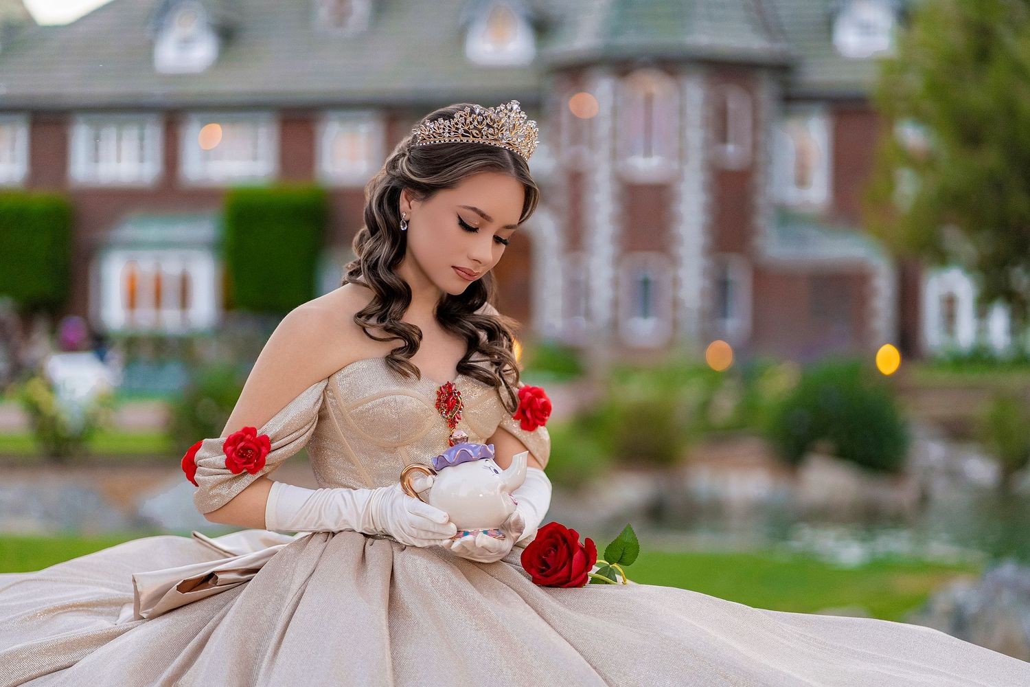 Young woman in elegant champagne ball gown and tiara holds red roses, posing regally before a grand manor house.