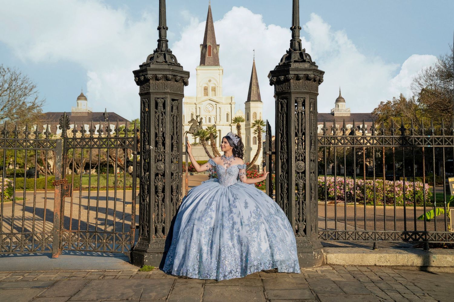 Young woman in blue quinceañera ball gown poses at ornate iron gates with St. Louis Cathedral in background.