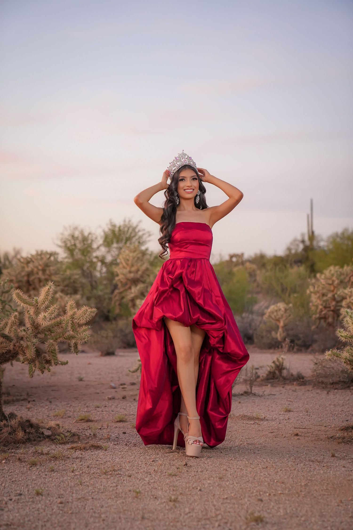 Young woman in red high-low gown and tiara poses in desert landscape with cacti at sunset.