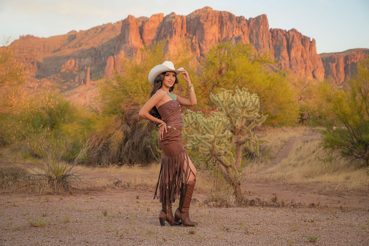 Woman in brown fringe dress and white cowboy hat poses before Superstition Mountains at golden hour in Arizona desert.