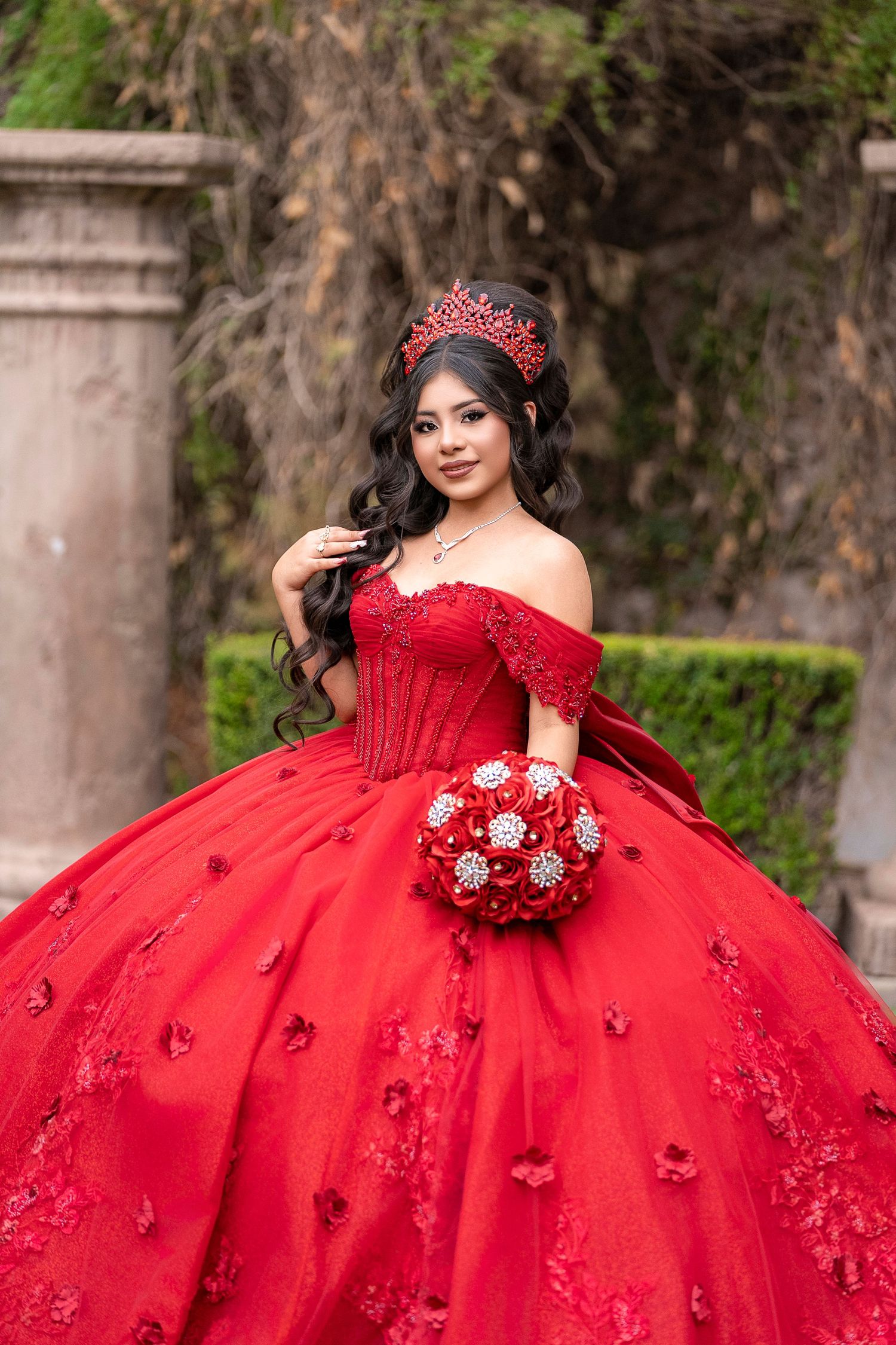 Beautiful young woman in elaborate red ball gown quinceañera dress with crown holds decorative red and silver bouquet.